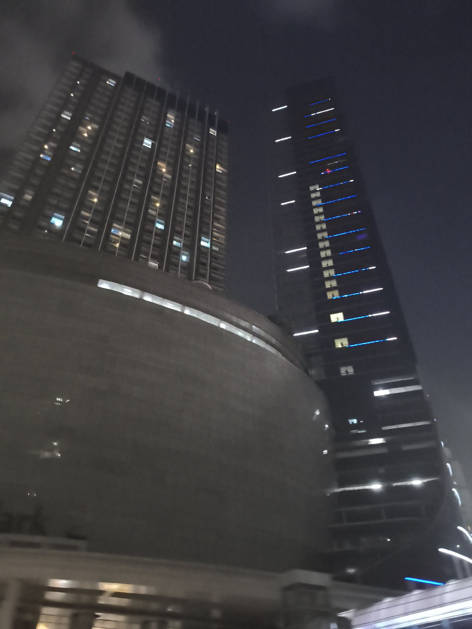 The image captures a nighttime view of two modern skyscrapers in West Jakarta, Indonesia. The skyscraper on the left is a tall, rectangular building with numerous windows, some of which are illuminated, suggesting occupancy. It appears to be older and has a more uniform facade. In contrast, the skyscraper on the right is sleeker and more contemporary, featuring a dark glass exterior adorned with striking blue and white LED lights that trace vertical lines up its sides. The lower portion of the image is dominated by a large, curved structure, possibly a podium or an adjacent building, which is softly lit and reflects some of the surrounding lights. The sky is dark and slightly hazy, with visible clouds, contributing to the urban nighttime atmosphere. The overall scene depicts a dynamic city landscape with architectural contrasts between older and newer structures.