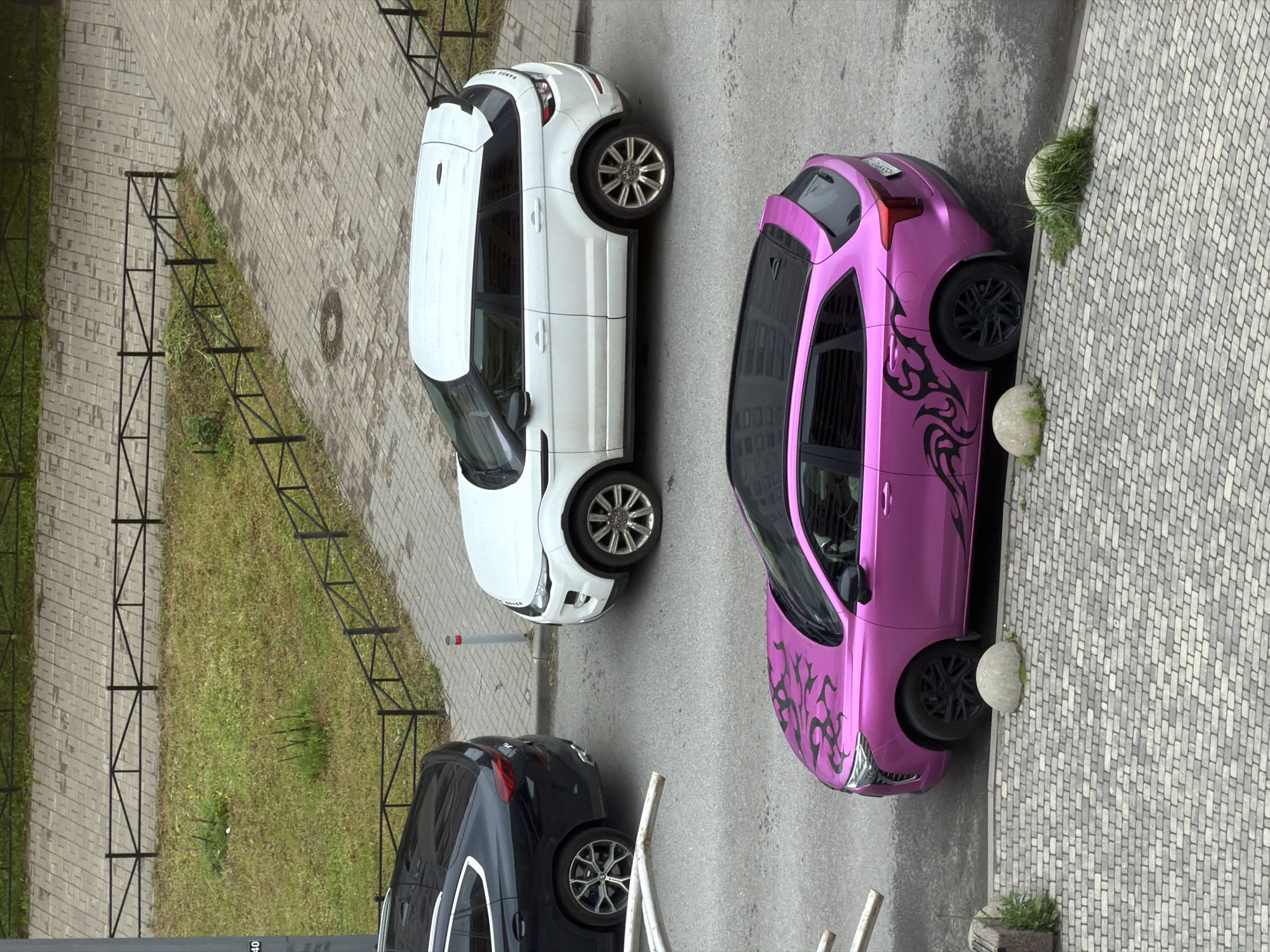A high-angle medium shot captures three parked cars on a street in Saint Petersburg, Russia. In the foreground, a vibrant pink SUV with black tribal decals on its hood and sides is parked on asphalt next to a cobblestone sidewalk bordered by two rounded concrete bollards with patches of grass growing around them. Behind the pink SUV, a white Range Rover Evoque is parked. To the left of the white SUV, a dark-colored BMW X6 is parked. The street is bordered by a grassy area with a black metal fence and a paved walkway. The scene appears to be on a cloudy day, indicated by the diffused lighting and lack of strong shadows.