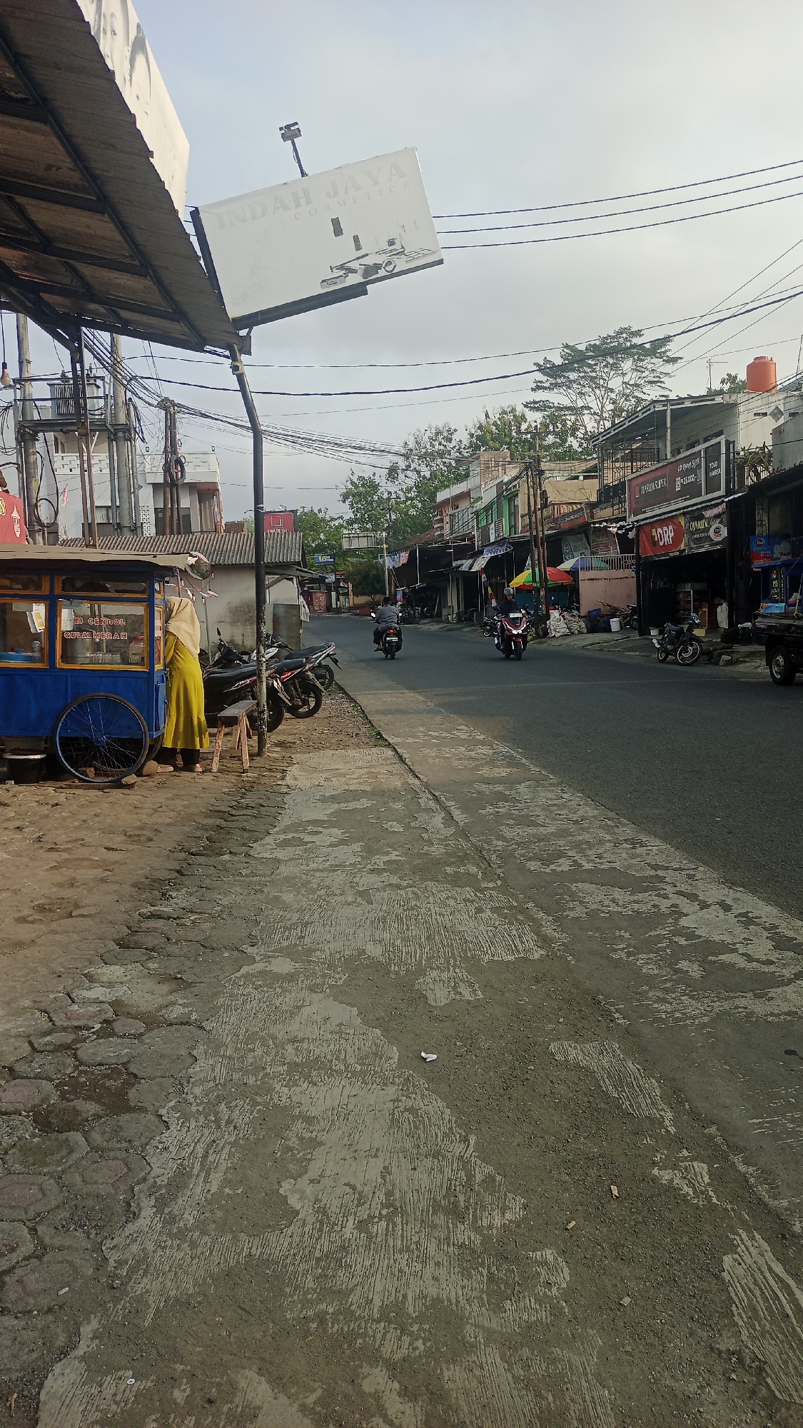 The image shows a street scene in Karyamandala, Indonesia. On the left side of the street, a food cart with a blue body and yellow trim is parked, with a woman in a yellow hijab standing next to it. Several motorcycles are parked on the sidewalk. A sign above the cart reads "INDAH JAYA COSMETICS". The street is paved and appears to be a busy thoroughfare, with several shops and buildings lining the right side. Motorcyclists are seen riding down the street. The sky is overcast.