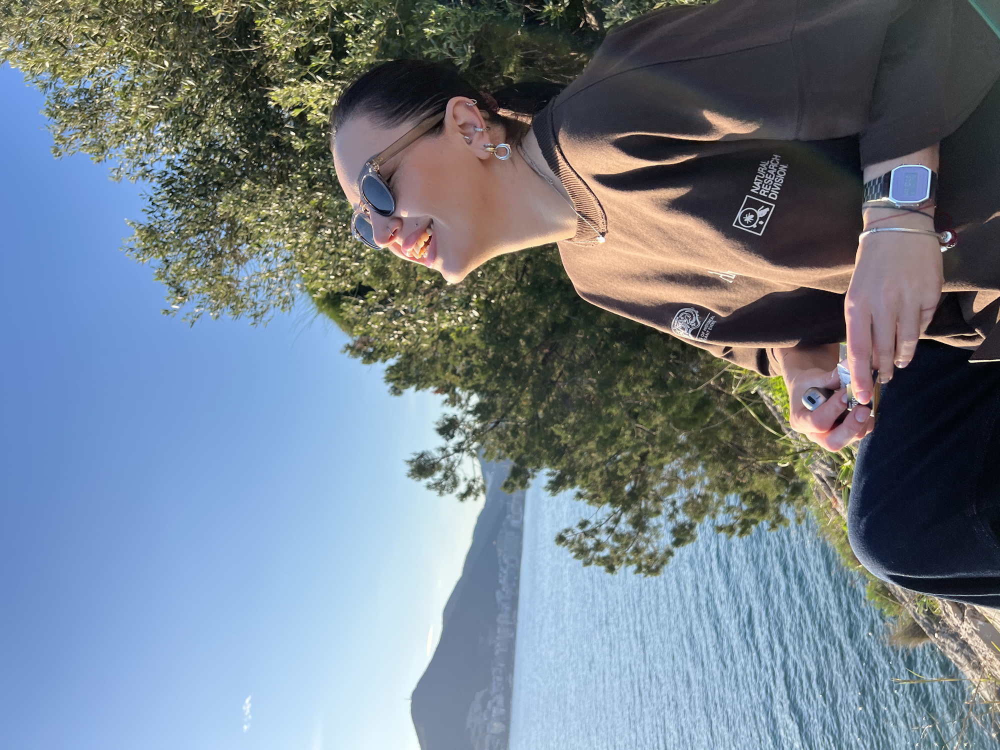 A woman in sunglasses and a brown "Natural Research Division" sweatshirt is visible in profile, looking off-frame towards a body of water. She is sitting outside near trees. The background shows a landscape including the sea, a mountain, and the city of Budva, Montenegro.