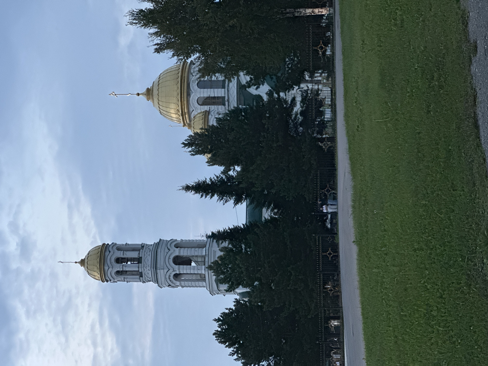 The image shows a view of the Transfiguration Cathedral in Lysva, Russia. The cathedral features white walls, green roofs, and a prominent golden dome, as well as a tall bell tower topped with a golden dome and a cross. Several tall evergreen trees partially obscure the building. In the foreground, a black metal fence with decorative gold elements runs along a paved road. A strip of green grass is visible in the lower part of the image. A few individuals are standing near the fence, appearing to be looking at the cathedral or interacting with each other. The sky is overcast with muted tones.