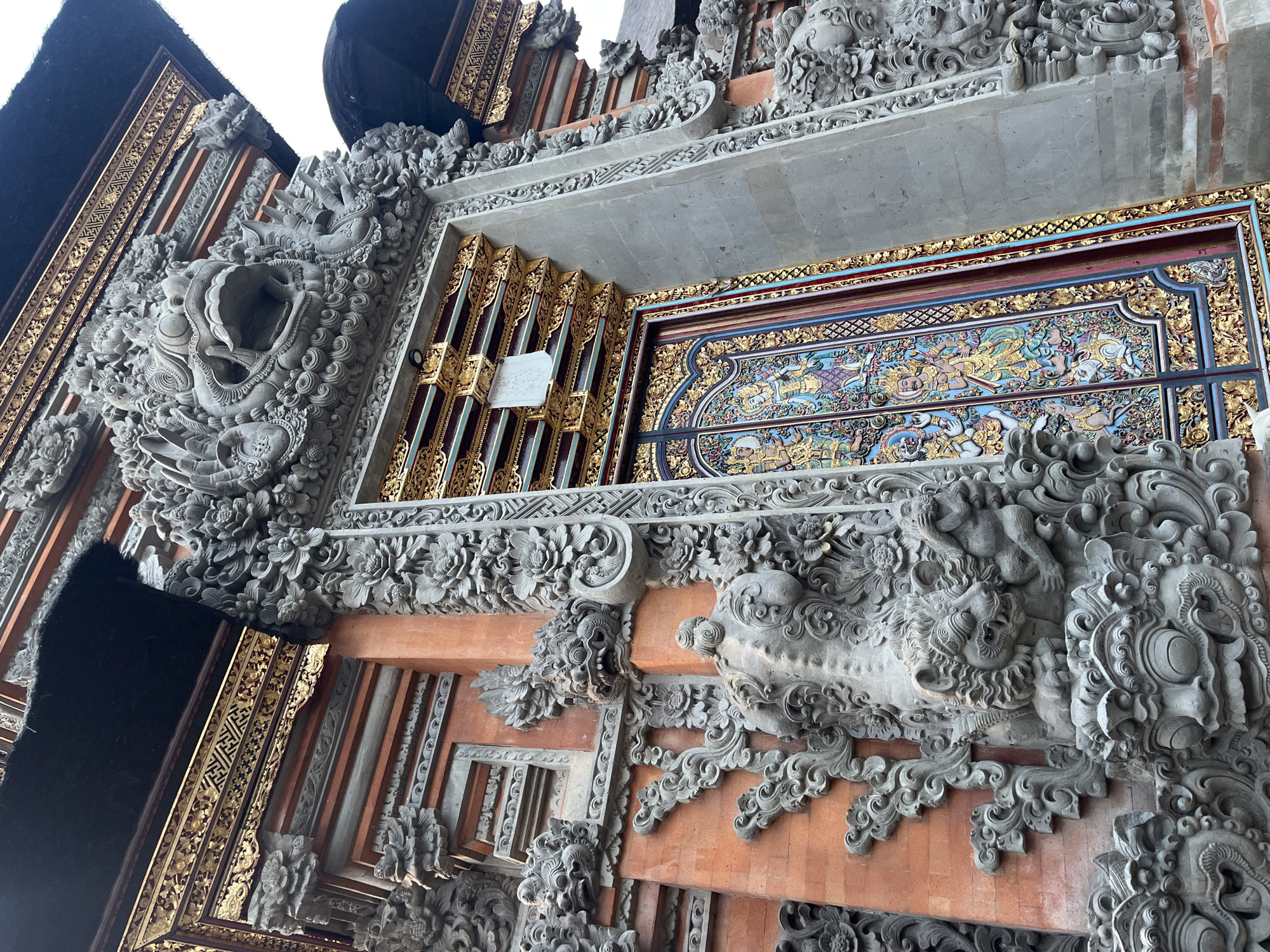 The image shows an ornate building structure in Ubud District, Indonesia. The exterior features intricate carvings in gray stone depicting floral patterns and mythological figures, contrasting against sections of exposed red brick. Some areas are adorned with gold-colored decorative panels. A painted panel displays a scene with multiple figures in gold, blue, and red colors. The image is taken from a low-angle perspective, highlighting the height and detail of the architecture.