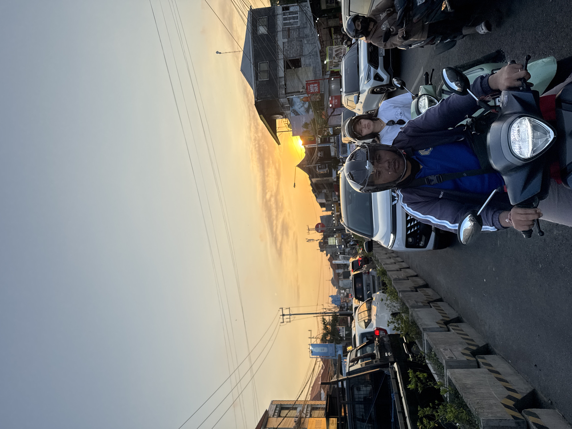 This is a vertical, eye-level, medium shot of a traffic jam during sunset on a city street in Denpasar, Indonesia. The sky is a gradient of pale blue to orange, indicating the sun is low on the horizon. Power lines crisscross the sky. The street is lined with buildings, some appearing to be shops or restaurants. A white SUV is in the foreground, followed by other cars and motorcycles. Several people are visible on motorcycles, wearing helmets. One person in the foreground is wearing a dark helmet and a blue shirt. Another person behind them is wearing a white shirt and a lighter helmet. To the right, another person on a motorcycle wears a brown hoodie and a helmet. Concrete planters with small green plants separate the traffic lane from the sidewalk or other areas. The overall scene depicts a typical urban sunset with traffic congestion.