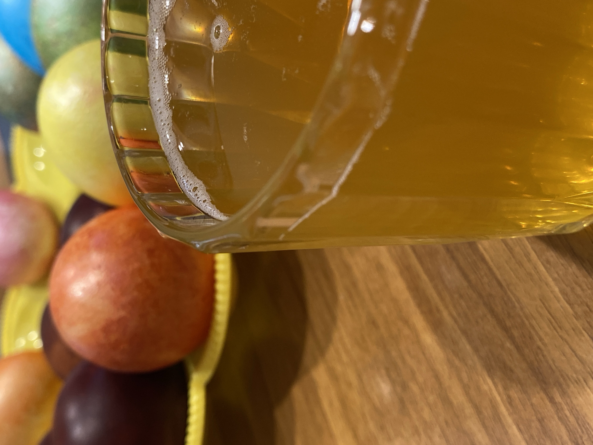 A glass mug filled with a yellow liquid, possibly beer, is positioned in the upper right quadrant of the frame, tilted at an angle. The mug's glass is clear and the liquid is bubbly. In the lower left quadrant, a plate of colorful candies is visible. The colors range from green, yellow, red, orange, and purple. The background is a wooden surface with a light brown color. The location and city cannot be determined from this picture. There is no indication to specify the location as Votkinsk, Russia.
