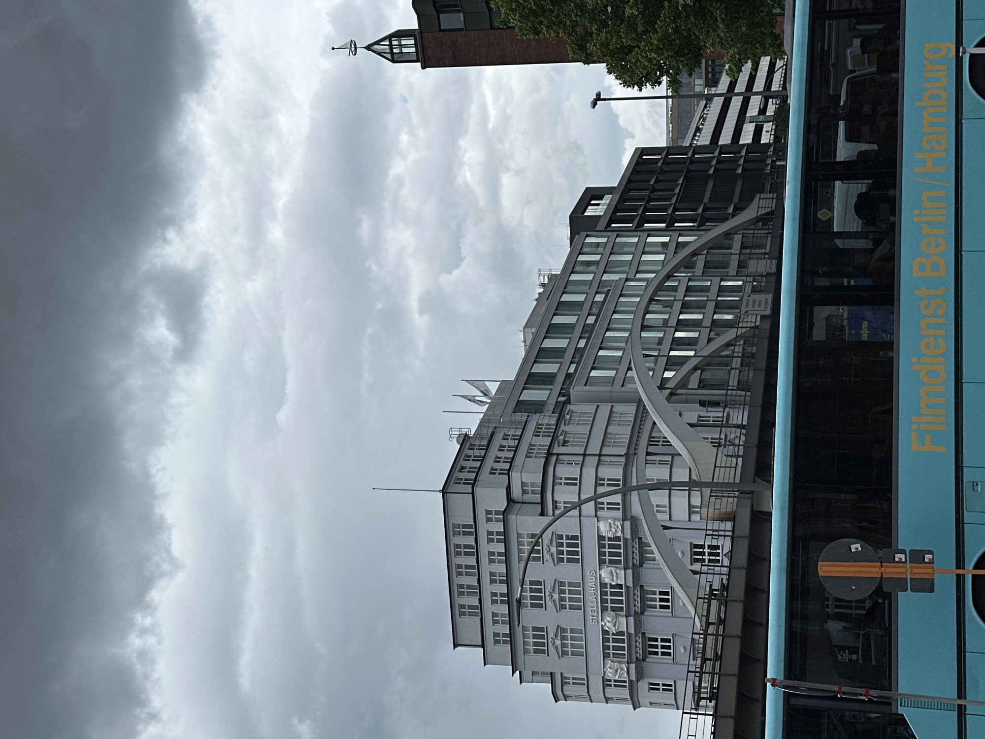 An urban scene in Hamburg, Germany, unfolds under a heavily overcast sky, filled with dense grey clouds suggesting a gloomy or pre-stormy day. The image, originally rotated, depicts a bustling cityscape.
Dominating the left side of the frame is the "STELLA-HAUS," a multi-story building with a light grey facade, featuring numerous square windows and decorative relief sculptures beneath some of them. The building has a distinctive stepped-back upper section. In front of it, a large, curved, silvery architectural structure sweeps across the mid-ground, possibly part of a pedestrian walkway or bridge.
Behind and to the right of the Stella-Haus, a more modern, dark-colored building with a grid-like window pattern is visible. Further back, a brick building with a striking pointed turret, topped with what appears to be a weather vane, rises against the cloudy backdrop. Green foliage from trees can be seen near these background buildings and above the bus.
On the right side, a light blue bus occupies a significant portion of the frame. Its side is visible, displaying reflections in its windows where silhouetted figures of passengers can be faintly discerned. Prominent golden text on the bus reads "Filmdienst Berlin/Hamburg." A circular street sign, yellow and black, is positioned near the front of the bus. The bus appears to be part of urban transit, indicating daily city activity. The heavy cloud cover suggests daytime, but not necessarily bright sunshine.