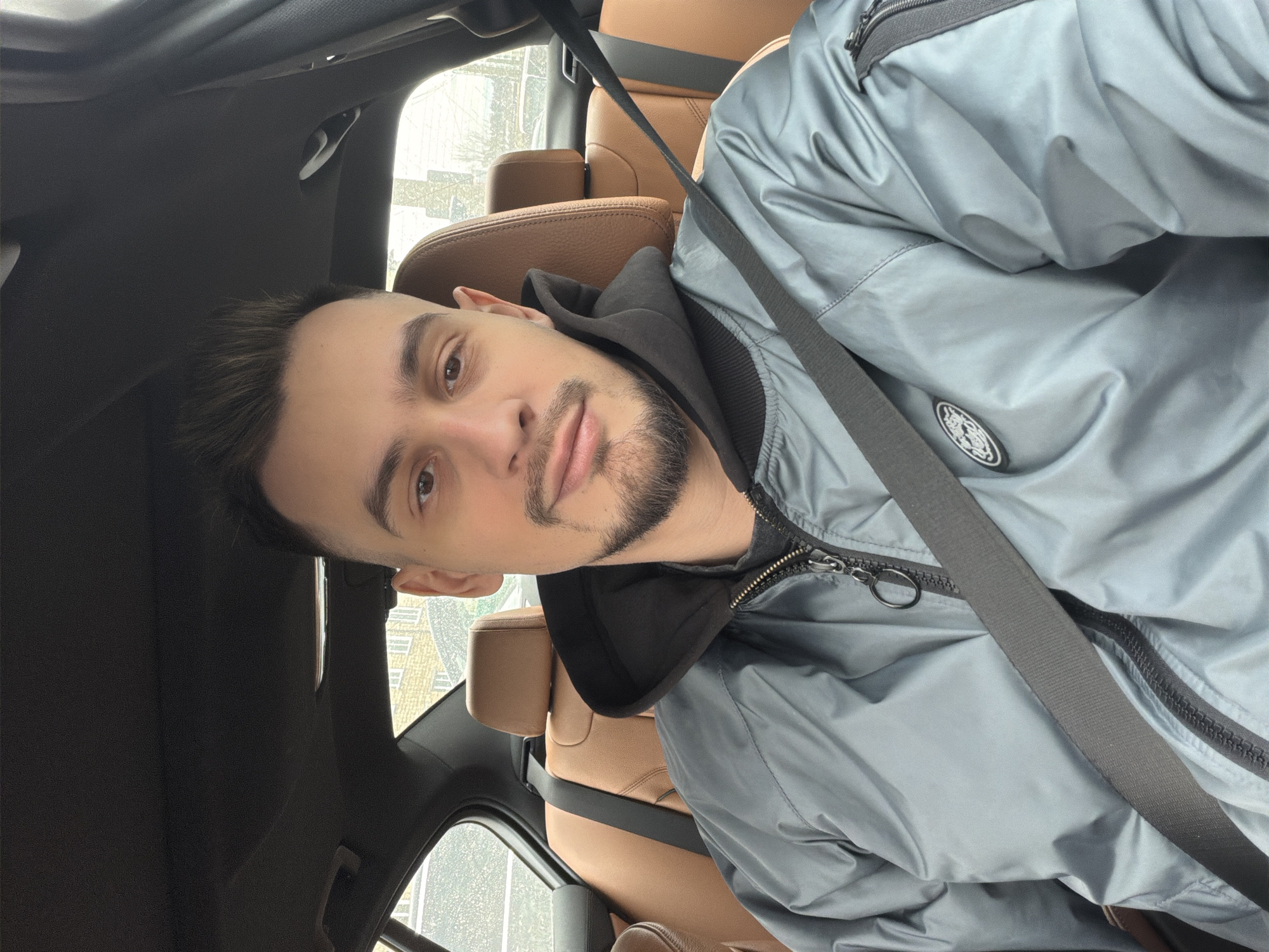 A male individual, estimated to be in his twenties to early thirties, is depicted in a close-up, chest-up view, seated within the interior of a vehicle, likely the driver's seat. The location is Krasnoyarsk, Russia. The subject has short, dark hair styled upwards, a short beard, and a mustache, and is looking directly into the camera with a neutral expression. He is wearing a black hooded sweatshirt beneath a light grey or silver bomber jacket. A black seatbelt is secured across his chest. The jacket features a front zipper with a metal pull ring and a zippered utility pocket on the left sleeve, below which is a circular patch. The vehicle's interior includes brown leather upholstery on the rear seats and front passenger headrest, and a dark headliner. Through the rear window, a light-colored brick building with multiple windows is partially visible, indicating an urban setting. The exterior weather conditions appear overcast or wet.