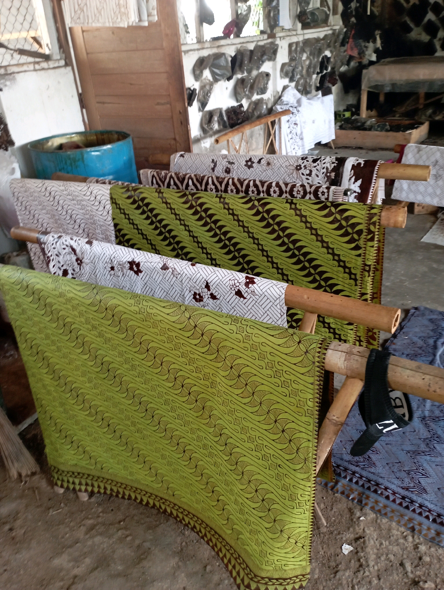 The image depicts a close-up view of a collection of batik fabrics displayed on wooden racks, likely in a workshop or market in Tasikmalaya, Indonesia. The fabrics are of various patterns and colors, with prominent green, white, and brown designs. Some fabrics have intricate geometric patterns, while others feature floral motifs. The racks are made of bamboo, and some are adorned with hanging items, including a black sock with white lettering. In the background, glimpses of a rustic interior can be seen, with wooden doors, a blue barrel, and walls adorned with various objects and materials. The overall impression is one of traditional textile craftsmanship.