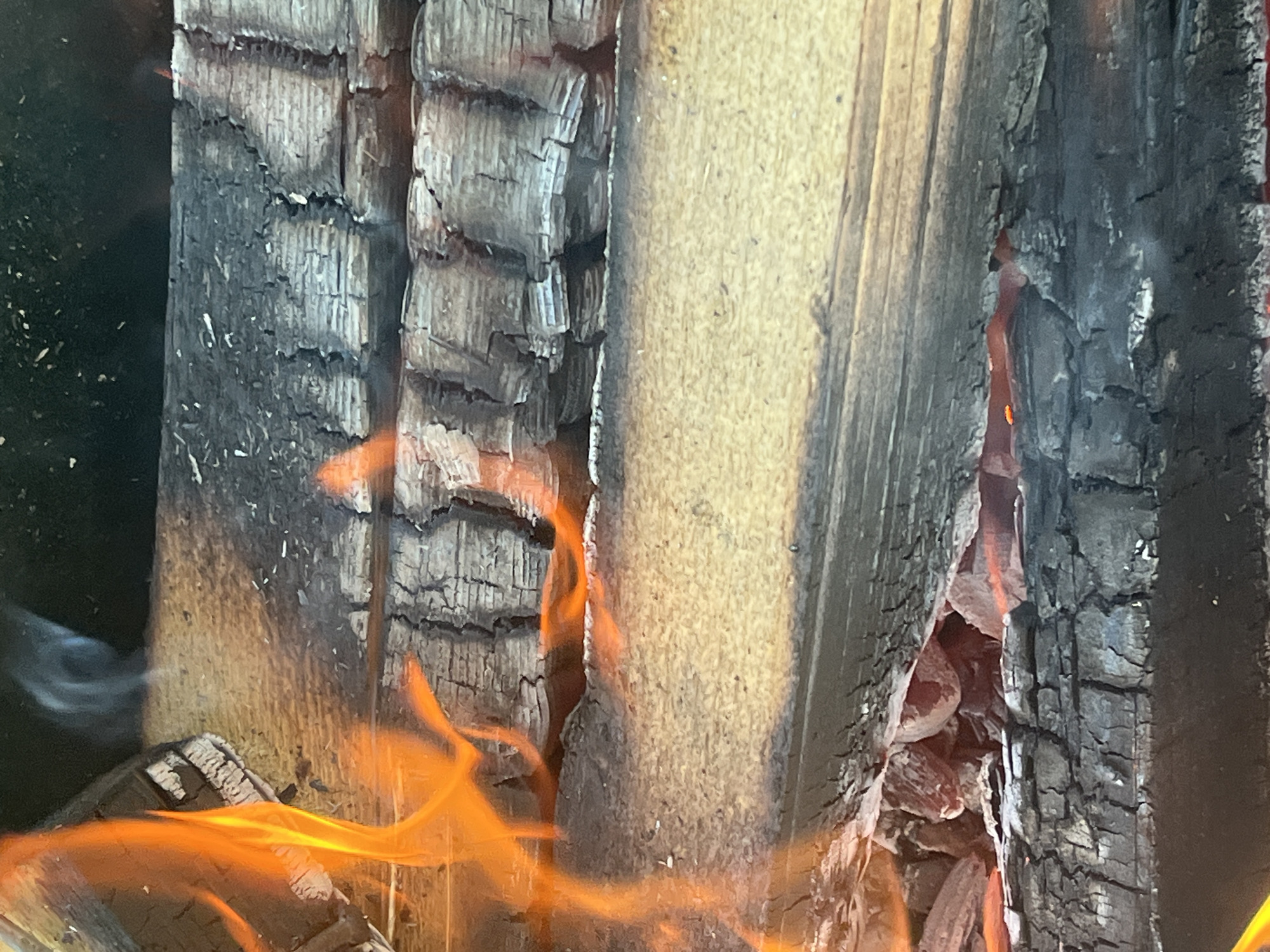 This is a close-up, overhead, eye-level photograph of burning logs in what appears to be a fireplace or fire pit. The image is dominated by textures and colors related to fire and wood.
In the foreground, a large, thick log is horizontally oriented, with its surface appearing charred and cracked. Flames, rendered in vibrant oranges and yellows, are actively consuming parts of the wood, particularly along the left side and in fissures within the logs. The fire casts a warm glow on the surrounding wood.
Above this prominent log, other logs are visible, also showing signs of burning. The upper surfaces of these logs are dark and ashy, with a textured, almost flaky appearance where the wood has been significantly affected by the heat. Thin wisps of smoke, appearing as faint grey and blue trails, rise from the burning areas and drift upwards into a dark, out-of-focus background.
The wood itself exhibits a rough grain and has undergone significant charring and splitting due to the intense heat. The lighting is dramatic, with the bright flames creating strong contrasts with the dark, burnt wood.
This photograph depicts a scene of active combustion of wood. The specific location in the city of Il'ino, Russia, cannot be determined from the visual information provided in the image.