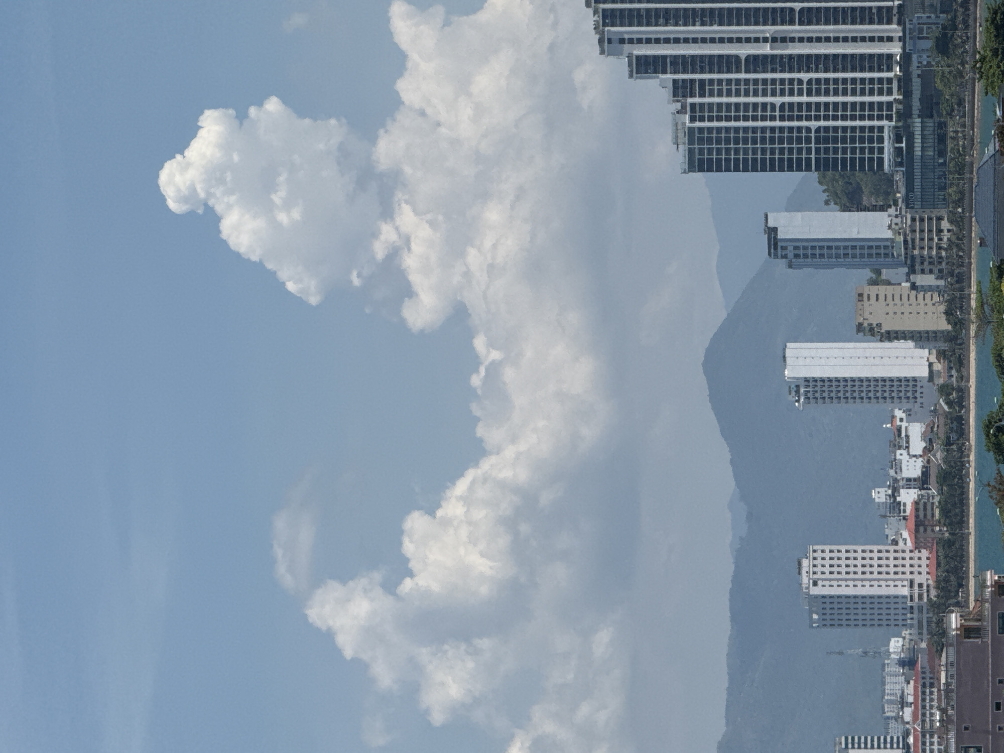 This is an image of a city skyline with mountains in the background. The sky is blue with large white clouds. In the foreground, there are several modern high-rise buildings. One building on the right is particularly tall and has a distinctive stepped facade. Other buildings are white and rectangular, with many windows. A body of water, likely the sea, is visible at the bottom of the frame, with some trees along its edge. The scene appears to be a coastal city.