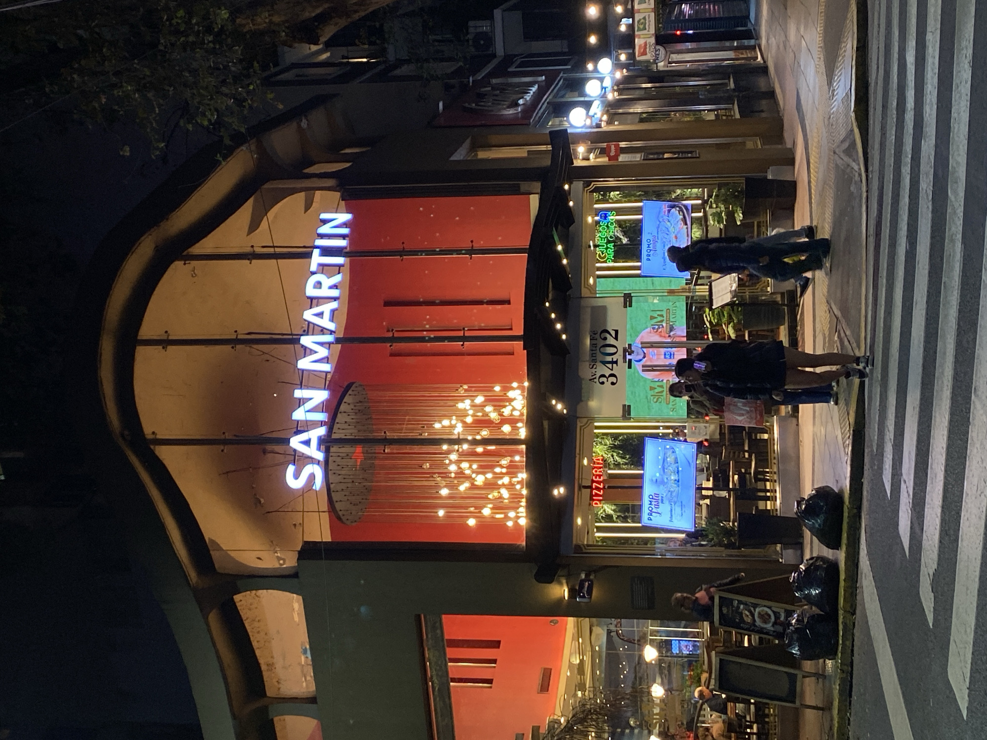 This night-time image captures a bustling street scene in Buenos Aires, Argentina, centered around a brightly lit commercial establishment. The primary subject is a restaurant or pizzeria named "SAN MARTIN," indicated by a large, glowing white neon sign prominently displayed on a red facade. Below the main sign, an intricate installation of small, warm-toned lights creates a decorative and inviting ambiance.
The establishment's entrance features large glass windows that offer a glimpse of the interior, showing seating and further lighting. A vertical sign on the right side of the entrance clearly displays the address: "Av. Santa Fé 3402." Two digital screens are visible in the windows; one advertises "PROMO Pasta" and another, featuring a cartoon character, reads "JUEGOS PARA CHICOS" (Games for Kids) and "PROMO Amigos." A smaller banner sign also identifies the place as a "PIZZERIA."
Several people are present on the sidewalk. Two individuals stand near a distinct black and white striped crosswalk, facing the establishment. Further along the sidewalk, another person is seen near a stack of black garbage bags placed on the curb. The scene is illuminated entirely by artificial light from the building's signage, interior lights, and streetlights, set against a dark night sky. Trees with dark foliage are partially visible in the upper left background. The image conveys an active urban evening environment.
