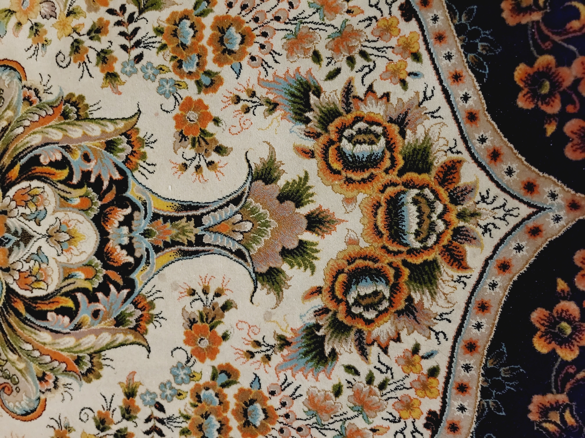 The image shows a close-up view of a highly ornate Persian rug. The rug features a complex floral pattern with vibrant colors, including shades of orange, blue, green, black, and cream. The central design appears to be a stylized tree or vase with elaborate scrolling leaves and flowers. Smaller floral motifs are scattered throughout the cream-colored background. The bottom of the image reveals a darker border with more intricate patterns and fringing. The rug is likely an antique or high-quality reproduction, indicative of a traditional aesthetic. Given the subject matter of a Persian rug, it is plausible that this item could be found in a museum, a private collection, or a traditional Azerbaijani home in Baku City, Azerbaijan. However, without further context or visual cues of the surrounding environment, it is impossible to definitively pinpoint its exact location within Baku City.
