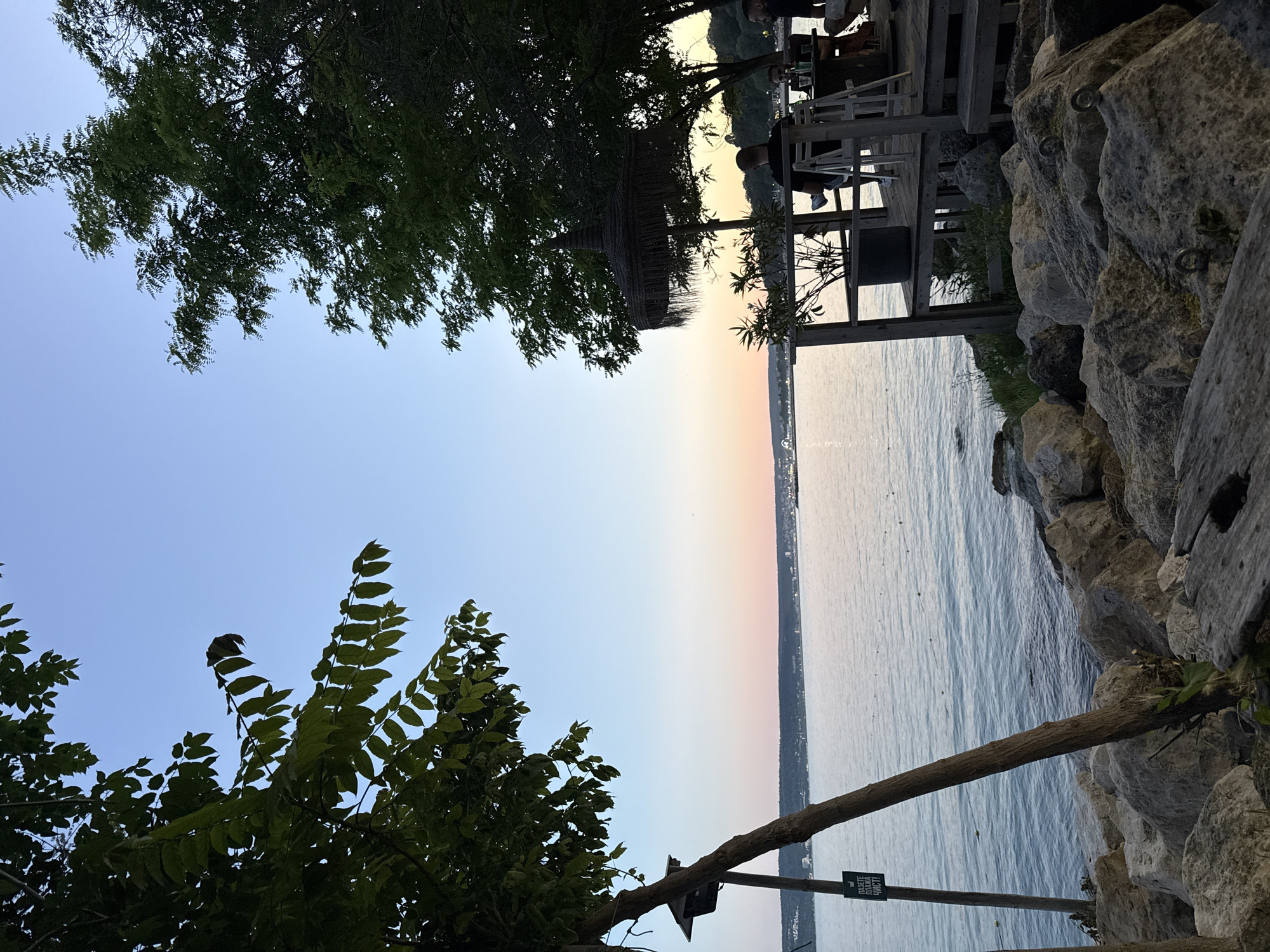 The image depicts a serene evening scene on the coast of Varna, Bulgaria, near the Sea Garden. The sky is a gradient of clear blue fading into soft pink and orange hues, indicating sunset. The tranquil sea stretches to the horizon, with a distant shoreline visible.
In the foreground, large rocks line the edge of the water, suggesting a natural, unmanicured beachfront. A wooden deck, part of a waterfront establishment, is situated to the right, featuring outdoor seating and a thatched umbrella. Several individuals are seated at tables on the deck, enjoying the ambiance.
Two trees frame the scene, one on the left with its branches reaching towards the sky, and another on the right providing shade over the deck area. A sign with Cyrillic text is attached to a pole near the left tree. The overall impression is one of a peaceful, relaxed evening by the sea.