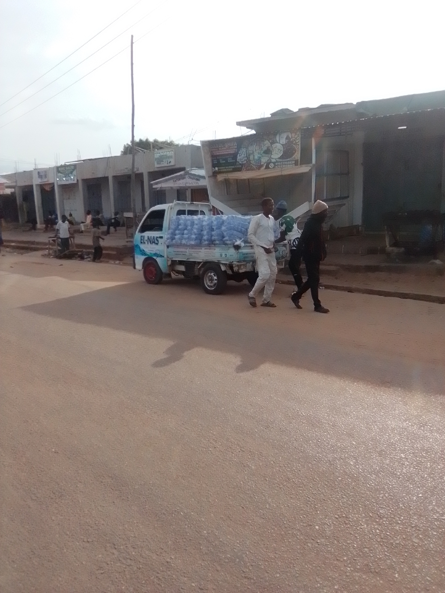 The image depicts a street scene in Galadima, Nigeria. A white flatbed truck is parked on the right side of the road, loaded with many plastic bottles of water. Three men are in the vicinity of the truck. Two men are walking away from the truck, one in a white outfit and the other in black. A third man, wearing a green and white shirt, appears to be near the truck's cargo. In the background, several buildings with storefronts are visible. The sky is overcast. The road surface is asphalt.