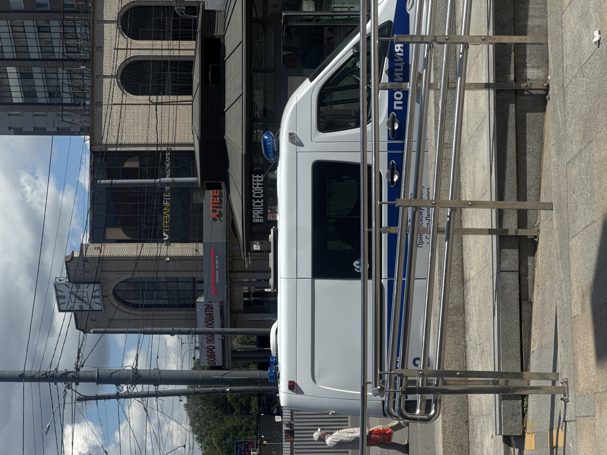 An outdoor, medium shot captures a white police van with blue flashing lights on its roof, parked beside a ramp with chrome railings. The van has the word "ПОЛИЦИЯ" (Police) in blue letters on its side. A person wearing a light-colored hat and jacket, carrying a red bag, walks away from the van and the ramp, moving towards the left side of the frame. The background reveals a street scene with buildings, electrical wires crisscrossing overhead, and a large clock face visible on one of the structures. A sign with Russian text, including "ДОБРО ПОЖАЛОВАТЬ!" (Welcome!), is also present. The environment appears to be an urban setting under a cloudy sky.