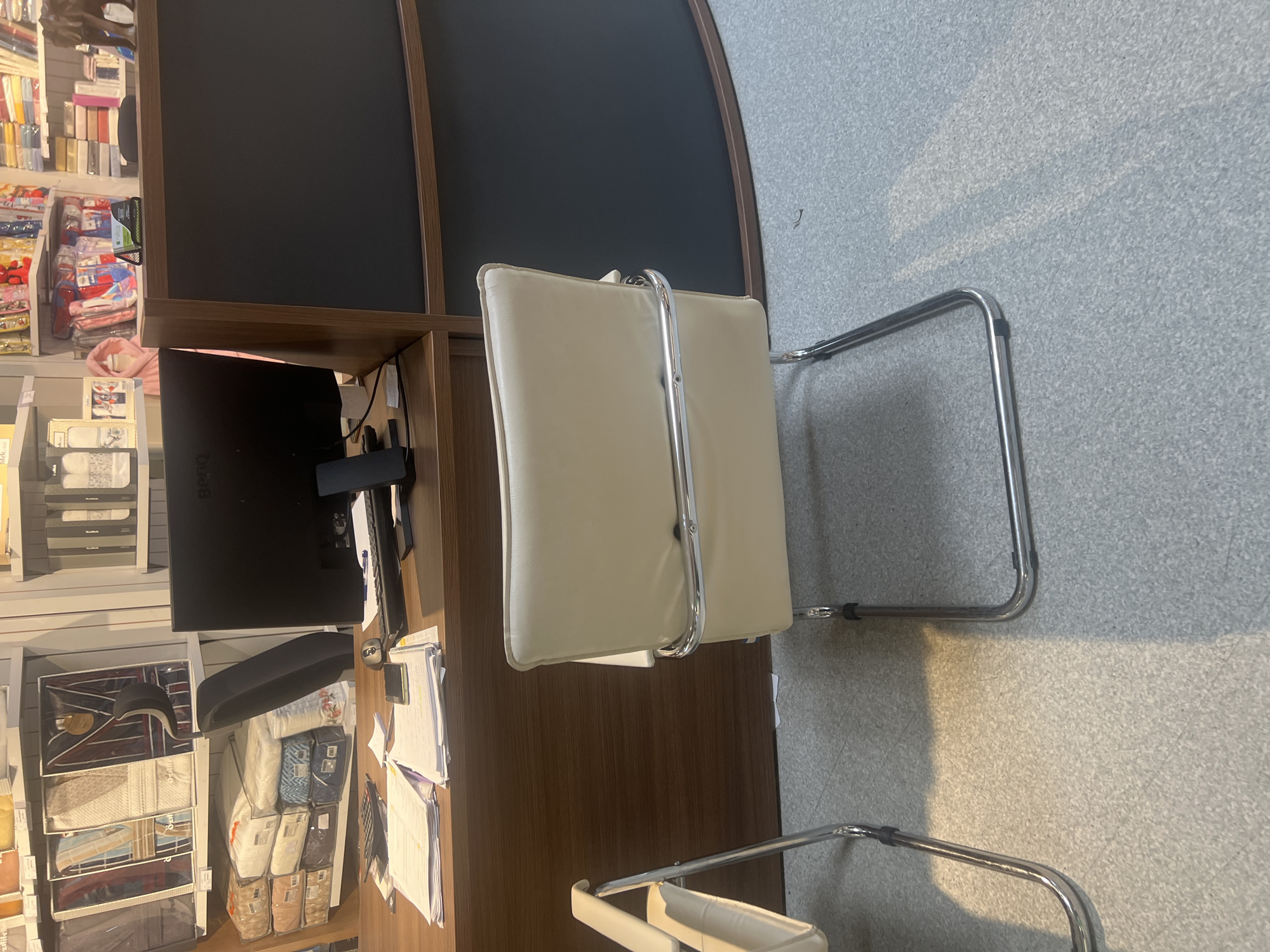 The image depicts a corner of a retail space, possibly a shop, featuring shelves stocked with various products, including textiles and bedding items. A desk or countertop is present, with a computer monitor, keyboard, and paperwork. Two beige office chairs with chrome frames are positioned near the desk. The environment includes a gray, carpeted floor. The location is unknown.