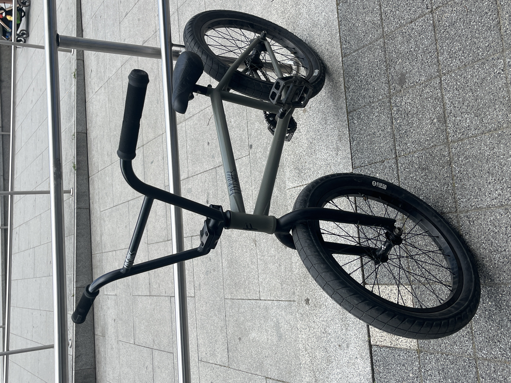 An olive drab-colored BMX bicycle with black components is positioned in a public urban setting, leaning against a polished metal railing. The bicycle features black handlebars with black grips, a black stem, and black front forks. Both front and rear wheels are black, equipped with black spokes and hubs. The tires are black with visible tread patterns; white lettering on the front tire sidewall reads "FUEGO," and "FIERO" is visible on the rear tire sidewall. A black seat, black pedals, and a black chain are present. A decal or sticker on the top tube of the frame, near the handlebars, partially displays "FIT BIKE" or "FMBK." The ground consists of grey square paved tiles with visible grout lines. In the background, beyond the railing, a blurred object resembling a baby carriage or stroller is partially visible. The scene is illuminated by even, diffuse light, casting minimal shadows. The location is in the city of Unknown, Unknown.