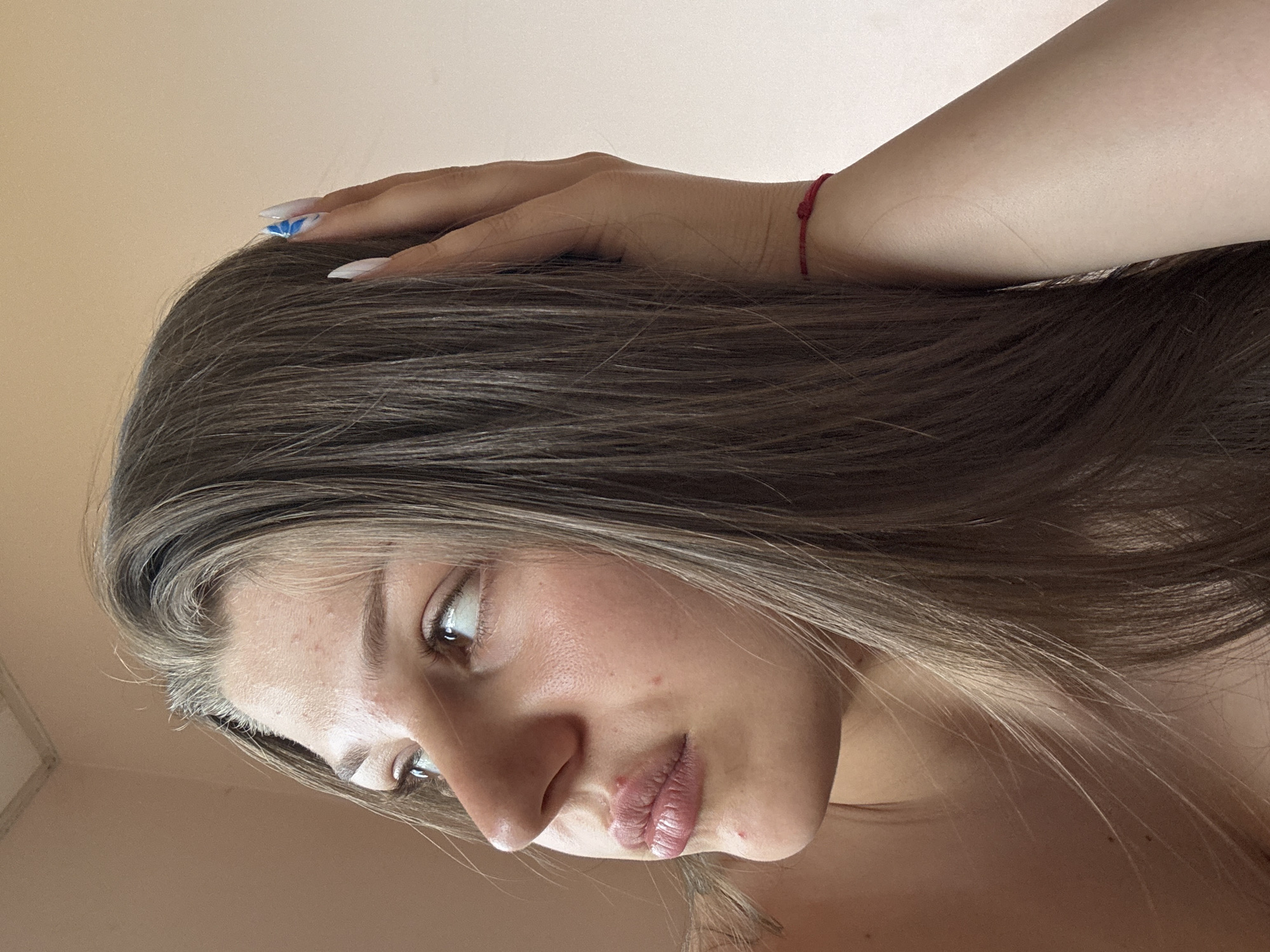 The image is an extreme close-up of a young person's face, tilted downwards and to the side. Their long, brown hair frames their face and extends into the foreground. The person has light skin with some blemishes visible. Their eyes are open and looking off to the left, with a slight hint of surprise or curiosity. Their lips are parted slightly, appearing to be glossy. A hand is resting on their head, with long, white fingernails that have blue tips, possibly nail art. A thin red string bracelet is visible on the person's wrist, peeking out from under their arm. The background is a plain, light brown color, suggesting an indoor setting. The lighting appears to be soft and natural, possibly from a window, creating subtle shadows on the face. No text is visible in the image. The overall mood is intimate and candid.