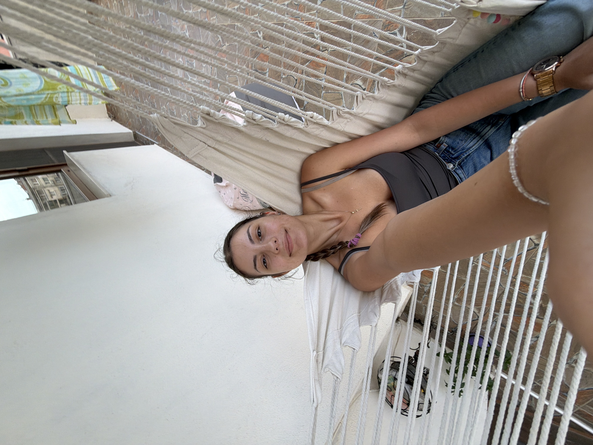A young woman with dark hair in a braid, wearing a dark grey tank top and blue jeans, is lying in a hammock on a balcony. She is holding up her left arm, extending it towards the camera. A thin, beaded bracelet is visible on her wrist. She is wearing a watch on her right wrist. Behind her, a white wall and some fabric draped over a railing are visible. The flooring of the balcony appears to be tiled. In the background, a window with a view of buildings and the sky can be seen. The scene is outdoors, likely on a balcony in the city of Tsarevo, Bulgaria.