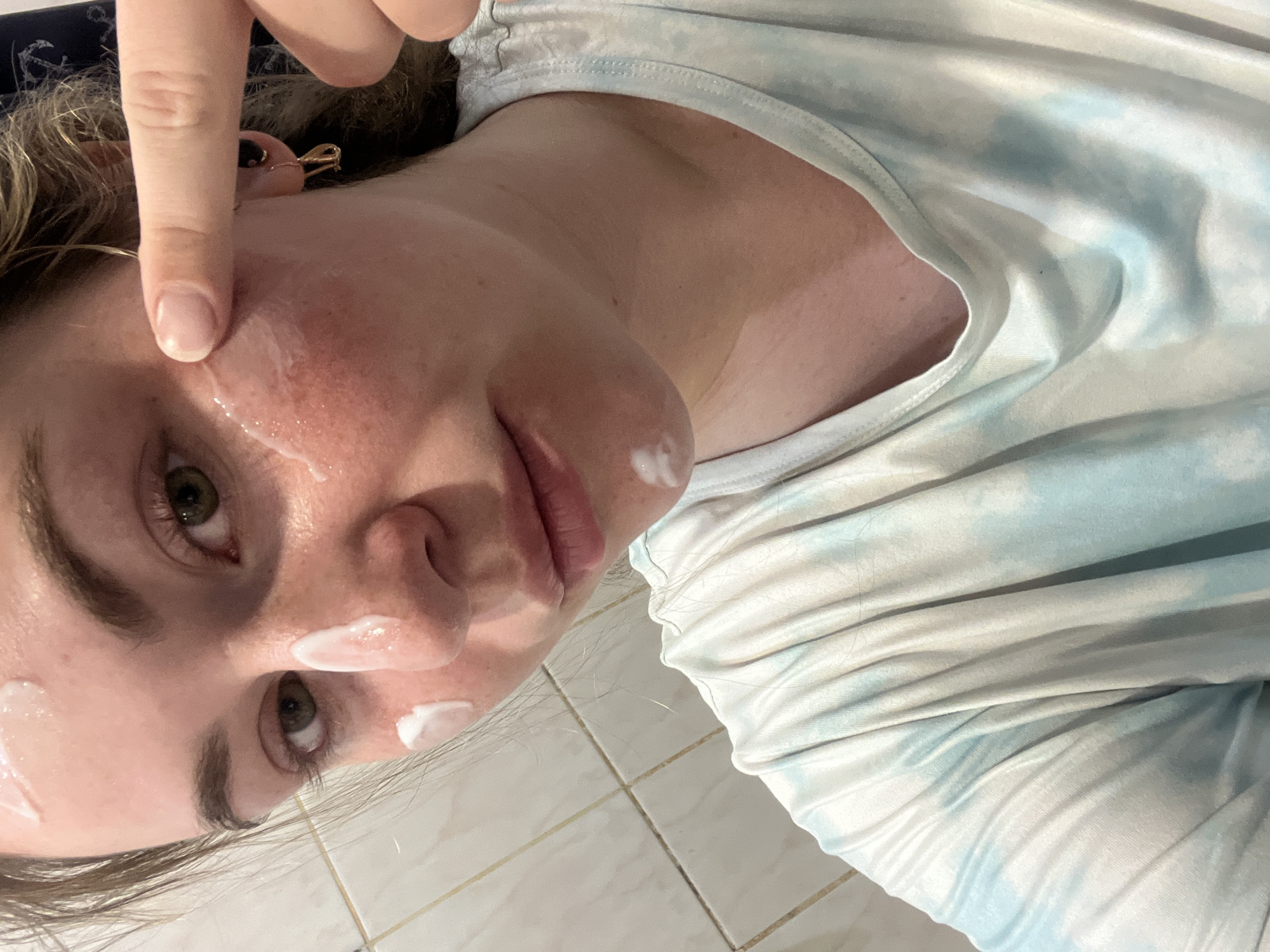 A selfie of a young, light-skinned person, presumably female, is oriented at an angle. The subject has fair skin, hazel eyes, and light-colored hair. White cream is visible on the face, specifically on the cheek, under the eye, and near the nose. A finger is pointed toward the cheek, likely in the process of applying or spreading the cream. The subject is wearing a light-colored t-shirt with a faded blue and white tie-dye pattern. The background includes beige tiles. The exact location in Chaikovsky, Russia, is not discernible from the visual information provided.