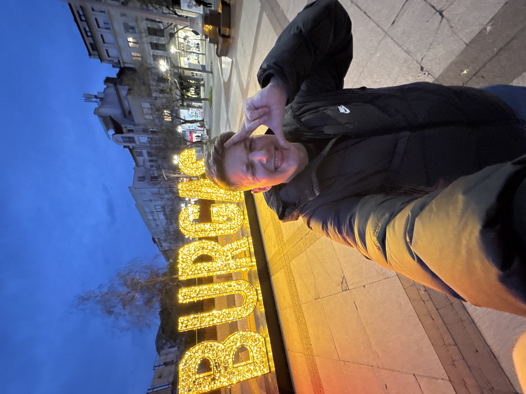 A young Caucasian male with short blonde hair, wearing a dark jacket with a Nike swoosh logo, is posing in front of illuminated yellow lettering that spells out "BURGAS" in a vertical arrangement. He is making a "V" sign with his fingers over his eye. The photo appears to be a selfie, taken at night. The background includes urban buildings, trees, and streetlights, consistent with a city center in Burgas, Bulgaria. The sky is a twilight blue. The ground is paved.