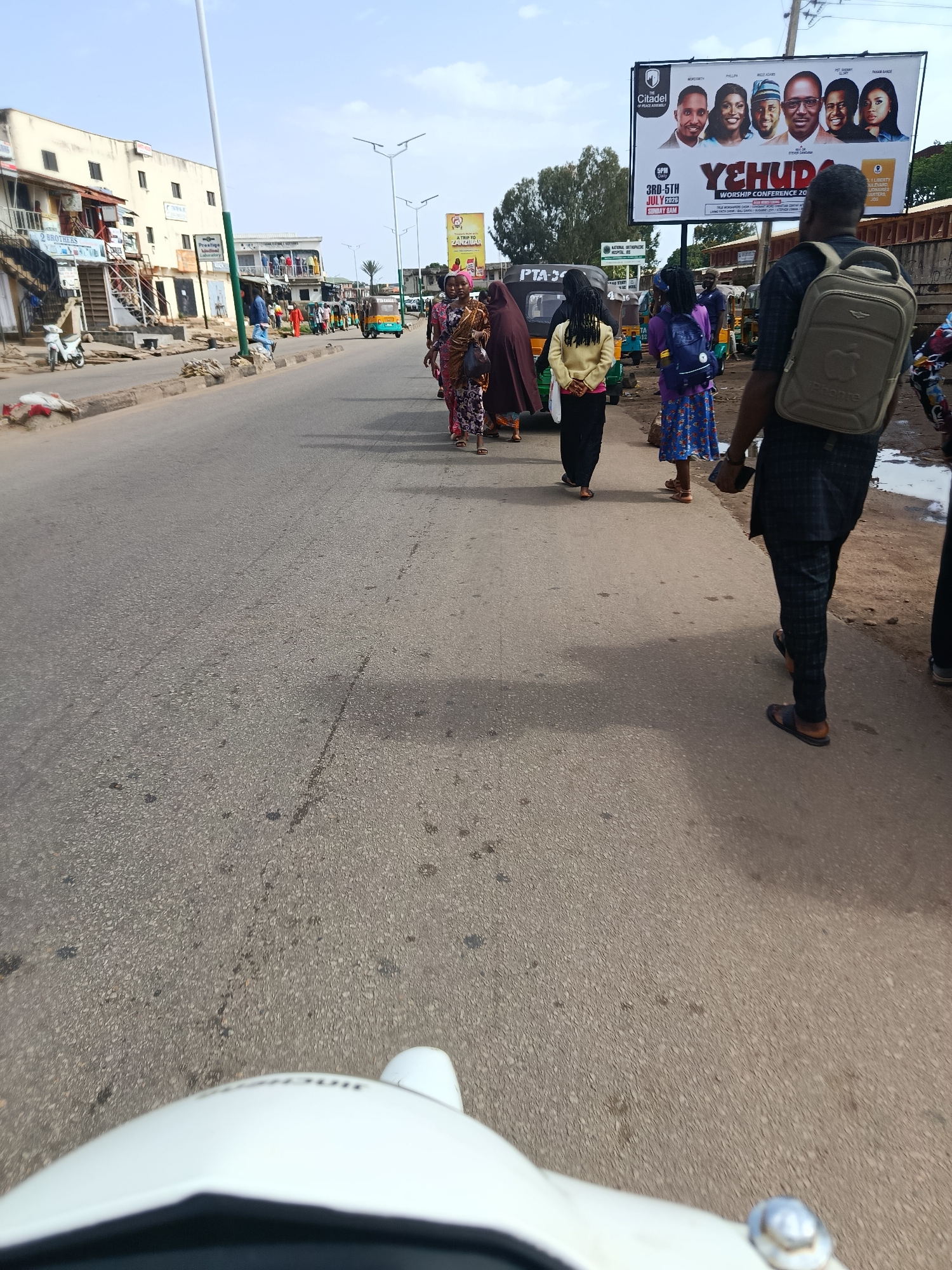 This media file captures a street scene in Jos, Nigeria, likely during the day given the bright lighting and clear sky. The image focuses on a road with several people walking along the side. In the foreground, a person is seen from behind, wearing a black and blue patterned outfit and carrying a beige backpack. Other individuals are visible further down the road, some walking in groups, dressed in various colorful traditional attire.
The setting appears to be a commercial or residential area, with buildings lining the left side of the street. One building has multiple levels with shops or offices, indicated by signage like "2 BROTHERS" and "Prestige Laboratory". Further down the road, a large billboard advertises a "YEHUDA WORSHIP CONFERENCE 2026" with dates and names of speakers. Several three-wheeled vehicles, commonly known as Keke Napep, are also present on the road, suggesting local transportation.
The weather appears to be clear and sunny. The overall atmosphere seems to be that of a bustling street with people going about their daily activities. The perspective suggests the photo was taken from a moving vehicle, possibly a motorcycle or a similar mode of transport, as indicated by the partial view of the vehicle in the bottom of the frame.