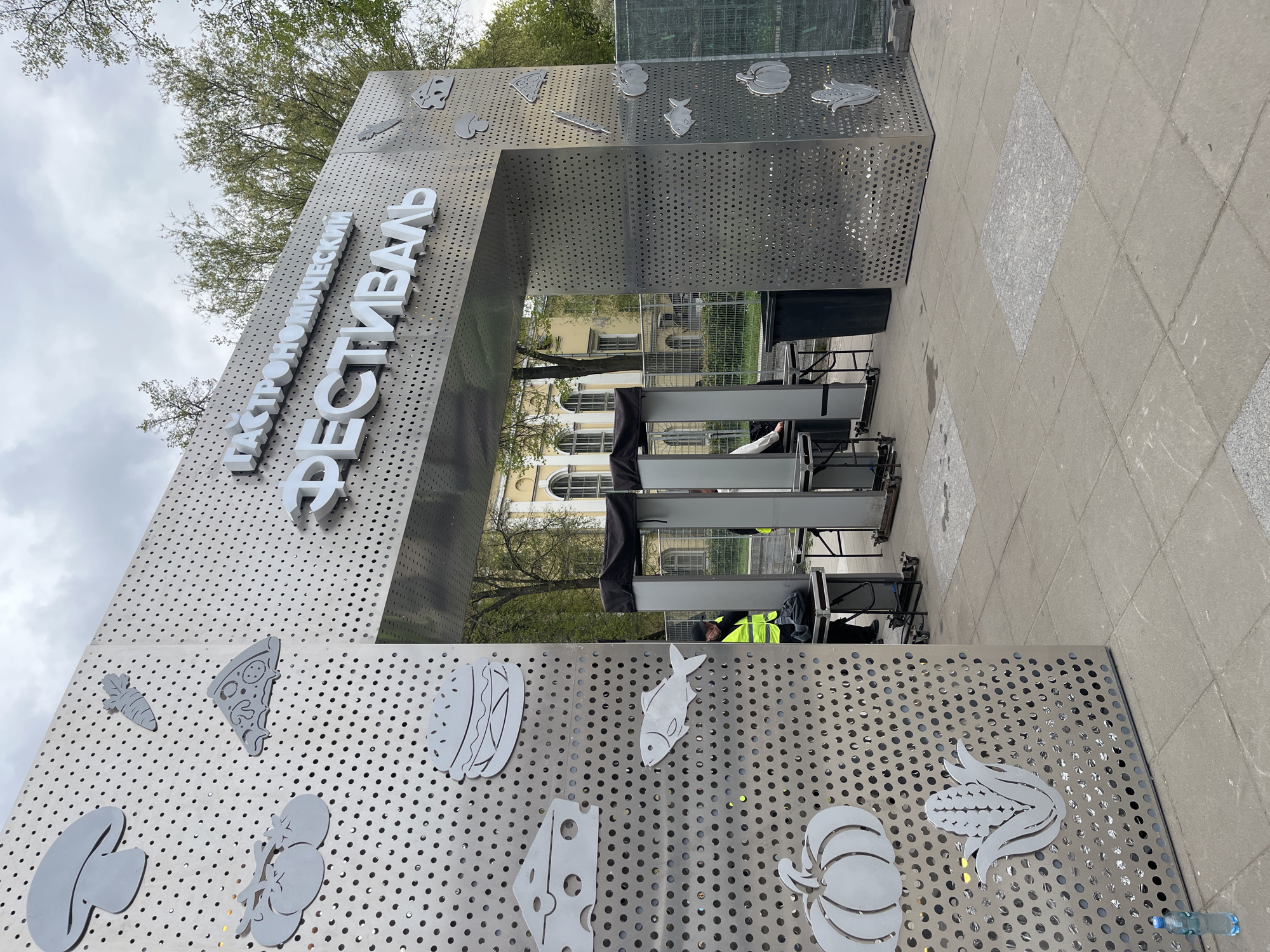 The image depicts the entrance to a culinary festival in Saint Petersburg, Russia. The entrance is a large, metal archway with a sign in Cyrillic that reads "Гастрономический Фестиваль" (Gastronomic Festival). The archway is decorated with metal cutouts of various food items, including a carrot, a pizza slice, a mushroom, a fish, corn, and a pumpkin. In the background, there is a building with white walls and arched windows. In the foreground, there is a security checkpoint with metal detectors. A person wearing a yellow safety vest is visible near the security checkpoint. The ground is paved with grey concrete tiles. The sky is overcast.