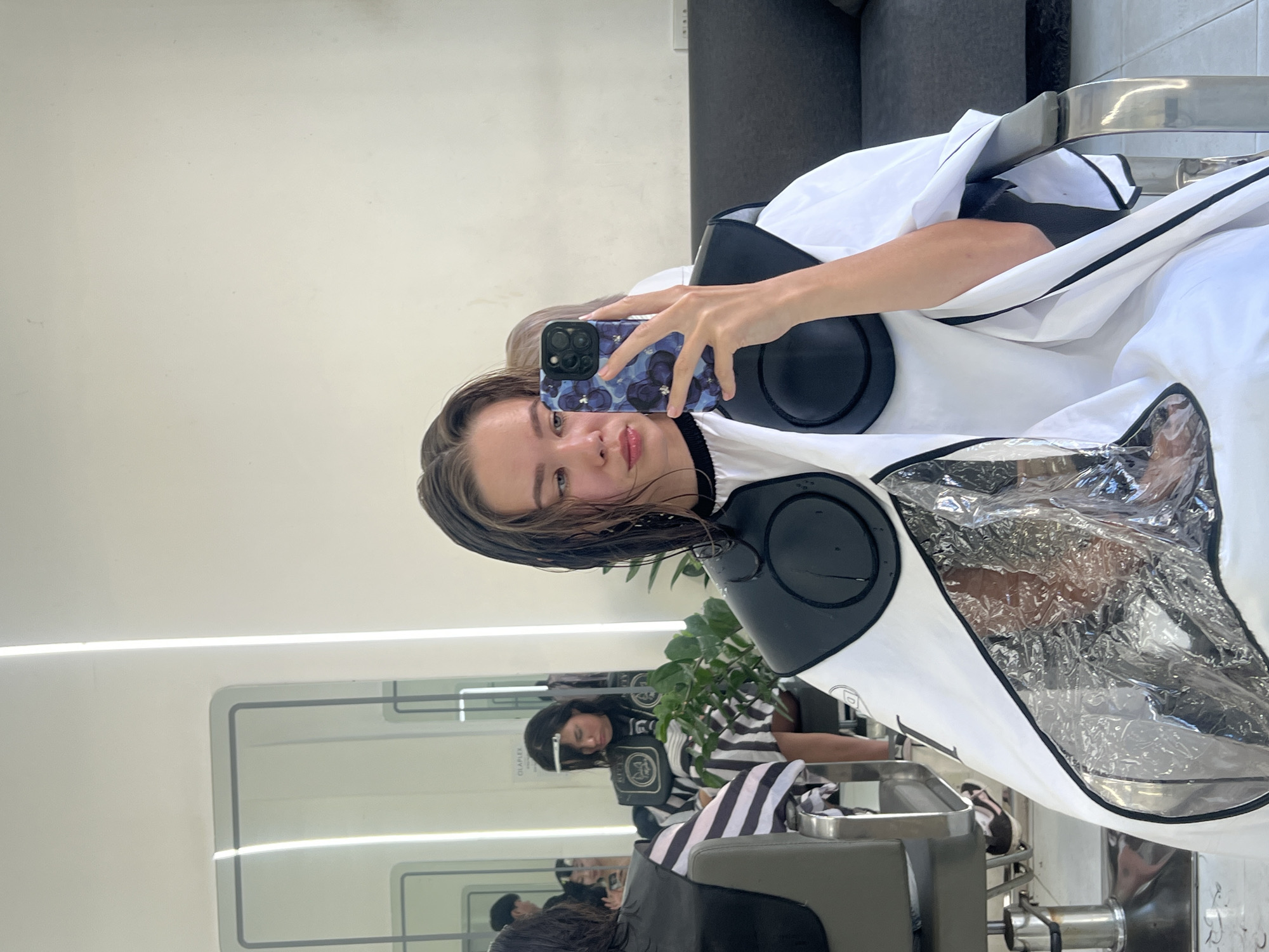A woman is in a salon, likely in the city of Nha Trang Ward, Vietnam, taking a selfie. She is seated and draped in a white salon cape with black trim, a clear plastic front, and black circular pads around the neck area. Her hair appears to be wet. She holds a phone with a blue floral case in her right hand. In the background, reflected in a mirror, another individual is also seated in a salon chair, wearing a similar salon cape. The environment suggests a modern salon interior with minimalist decor and adequate lighting.