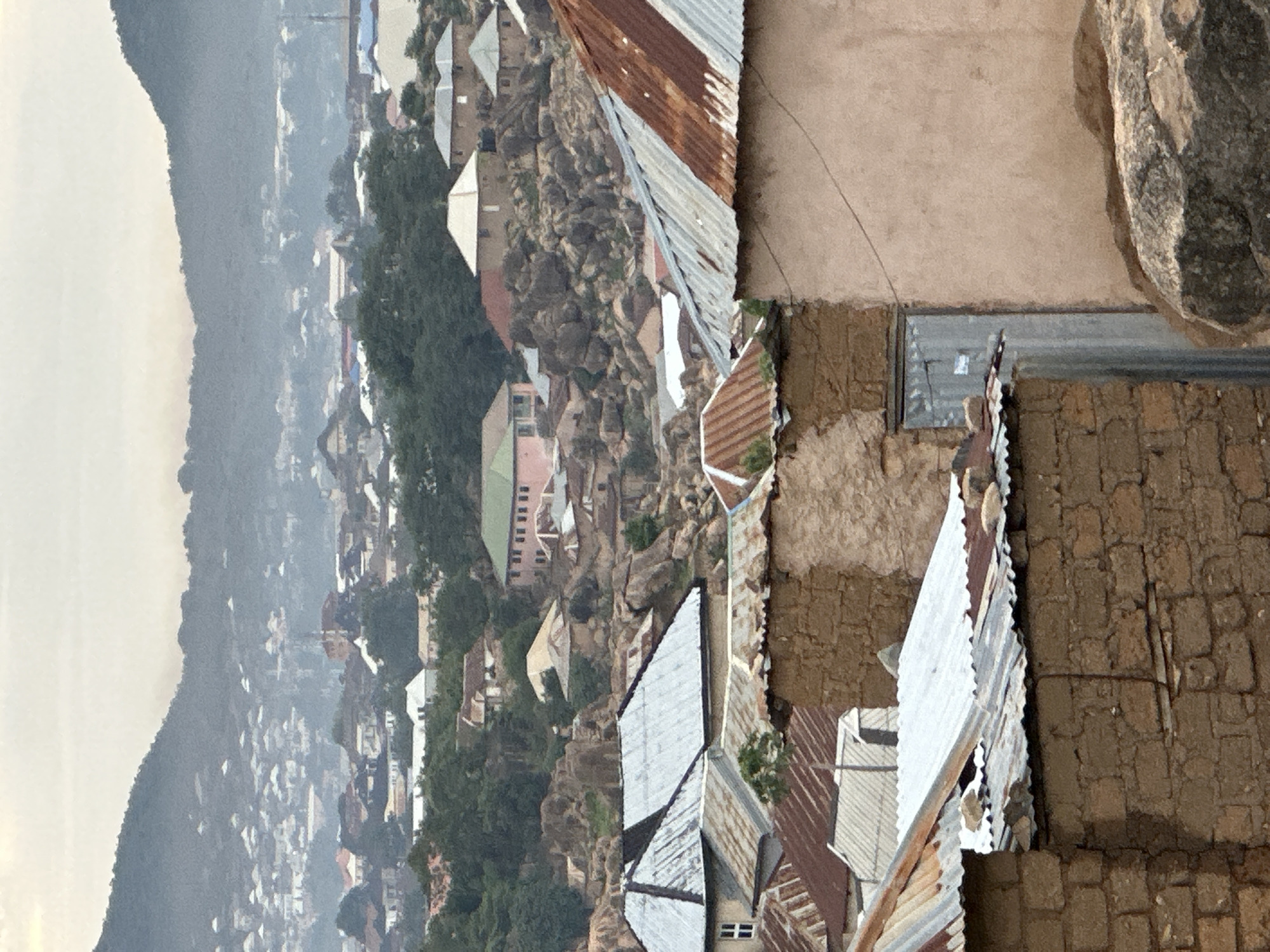 This image captures a panoramic view of a residential area in Jos, Nigeria, characterized by a rugged, rocky landscape interspersed with human settlements. The scene is dominated by hills in the background and numerous buildings extending into the midground and foreground.
In the immediate foreground, a cluster of buildings with various construction materials is visible. The closest structures feature walls made of rough-hewn, reddish-brown bricks or mud blocks, topped with corrugated iron roofs. Some roofs exhibit significant rust and wear, while others appear newer and silver. A narrow, corrugated metal door is visible on one of the foreground buildings. Large, weathered rocks are present around these structures, indicating the natural terrain.
The midground reveals a dense arrangement of residential buildings nestled amongst large, grey rock formations and scattered trees. Many of these houses also have corrugated iron roofs, varying in color from rusty brown to silver. Some buildings display plastered walls, including one prominent light pink or peach-colored multi-story structure with numerous small, dark windows. Another building in the midground has distinct white-framed windows. The terrain is notably uneven, with houses built on and around the boulders.
Further into the background, the urban area expands across a valley and up the slopes of distant hills. A thick atmospheric haze partially obscures the furthest structures, lending a muted blue-grey tone to the landscape. Numerous white-roofed buildings dot the distant hillsides, indicating a sprawling residential area. The silhouette of significant hills forms the horizon under an overcast or hazy sky. No people or active human interactions are discernible within the frame.