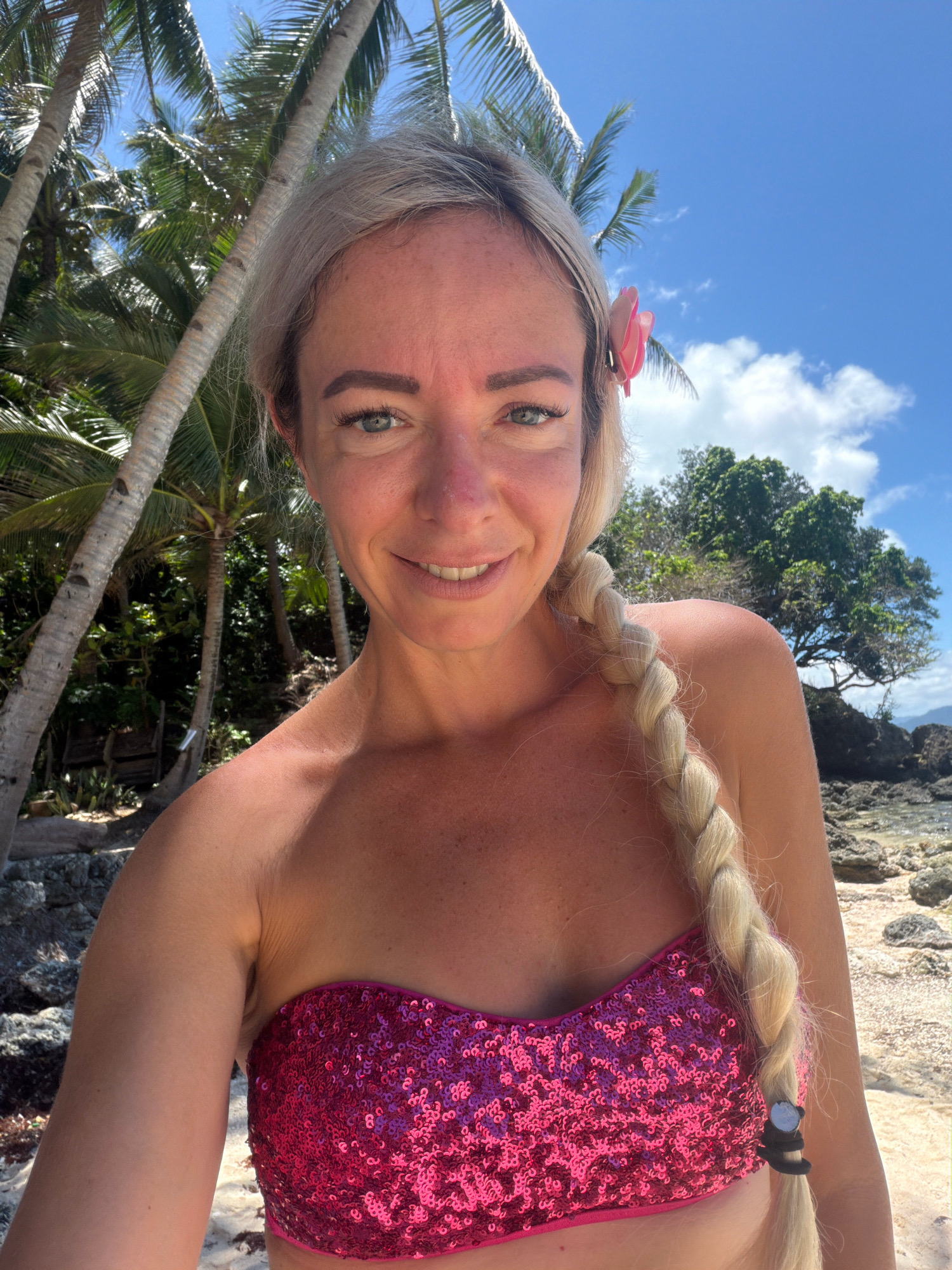 A Caucasian woman with blonde hair styled in a braid and a pink flower tucked behind her ear, smiles at the camera. She is wearing a sparkly pink bikini top and is standing on a beach with palm trees and a clear blue sky in the background. The location is the city of Malay, Philippines, likely on Boracay Island.