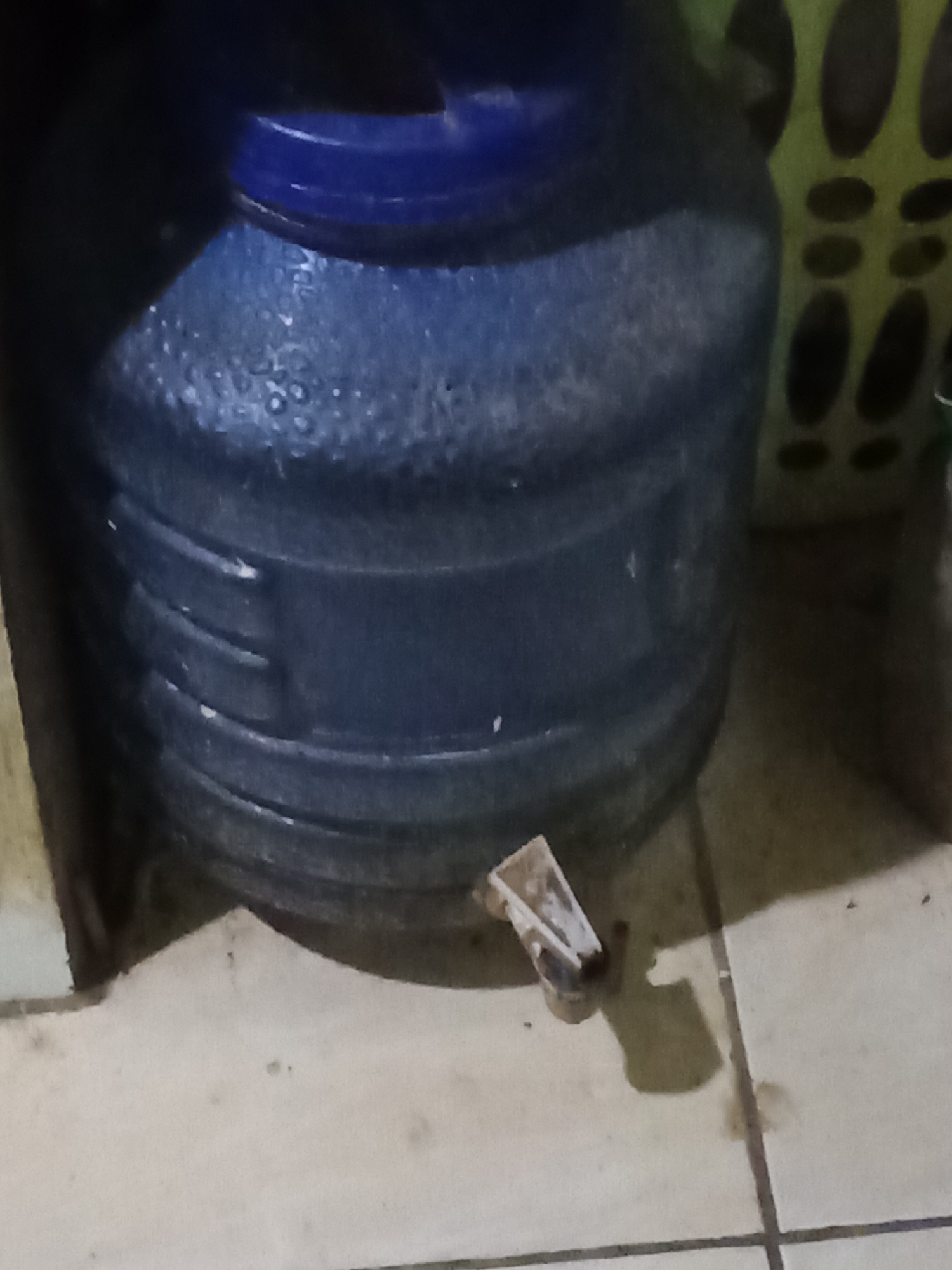 A close-up, horizontal shot captures a large, blue plastic water jug with a ribbed texture, positioned on a tiled floor. A small, detached plastic spigot lies on the floor in front of the jug, indicating a potential leak or a disassembled part. In the background, a portion of a green, perforated plastic container is visible. The lighting is dim, creating shadows and obscuring fine details. The scene appears to be an indoor environment, possibly a residential or commercial space in Pemalang, Indonesia. No individuals are present in the frame.