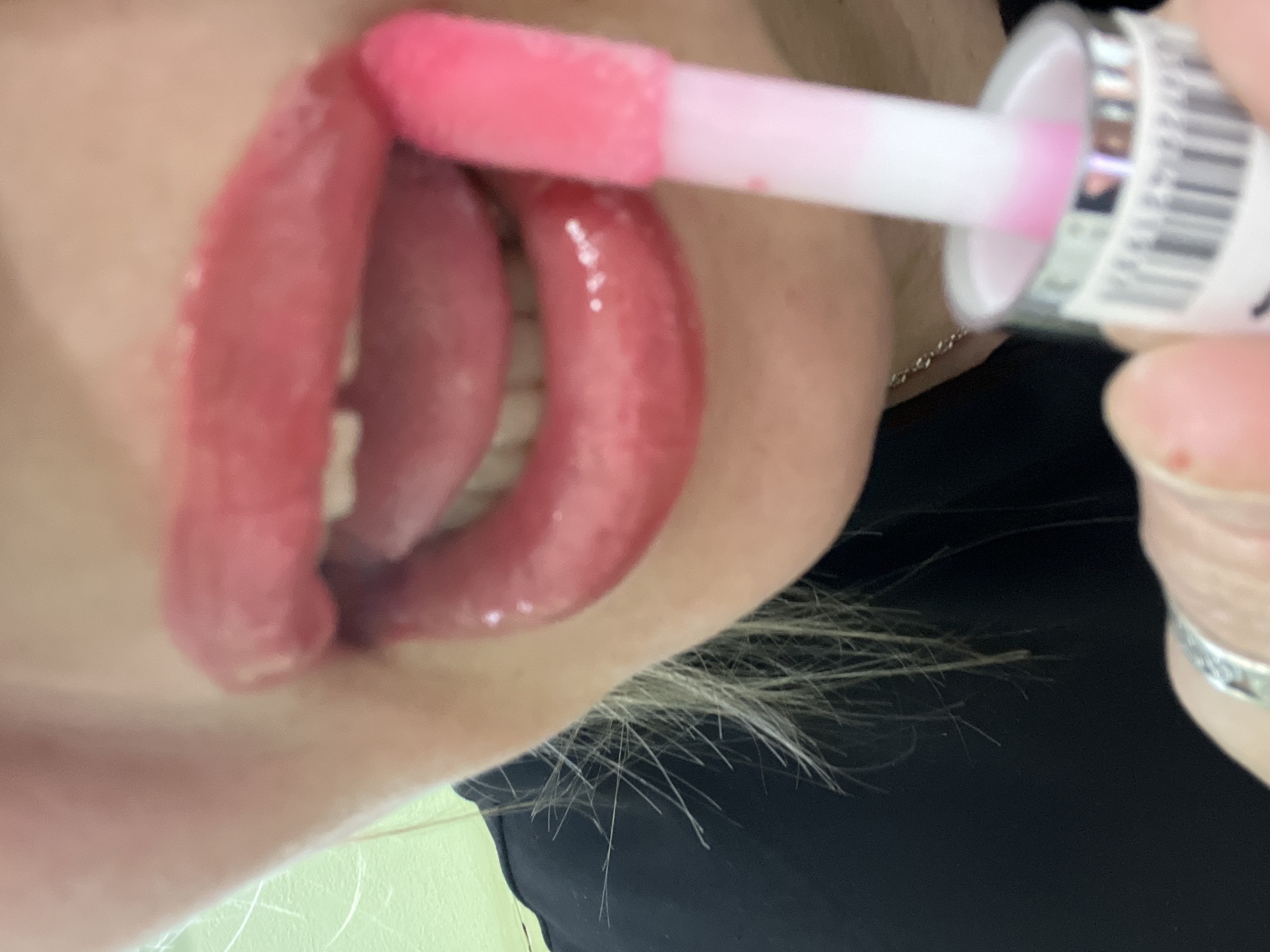 A close-up, angled shot captures a person in the process of applying pink lip gloss. The central focus is on the person's open mouth, where their lips are coated in a bright, shiny pink gloss, appearing plump and wet. A glimpse of white teeth is visible within the parted lips. A white lip gloss applicator, with a soft pink tip saturated with product, is held by a hand and touches the upper right side of the person's mouth, actively spreading the gloss. The hand, which has a light skin tone, wears a silver ring on one finger and holds the base of the lip gloss container. This container features a white label with blurry, indistinct black text or a barcode pattern.
The image is tightly framed, emphasizing the cosmetic application. Light-colored, wispy hair is visible along the bottom-left edge of the frame, hinting at the person's appearance. The background is softly blurred, showing areas of light skin, dark fabric (likely clothing), and some indistinct light green or beige surfaces, suggesting an indoor setting. The time of day is not discernible, and no specific emotions are conveyed. This activity takes place in Chaikovsky, Russia, though no explicit geographical cues are visible within the image itself.