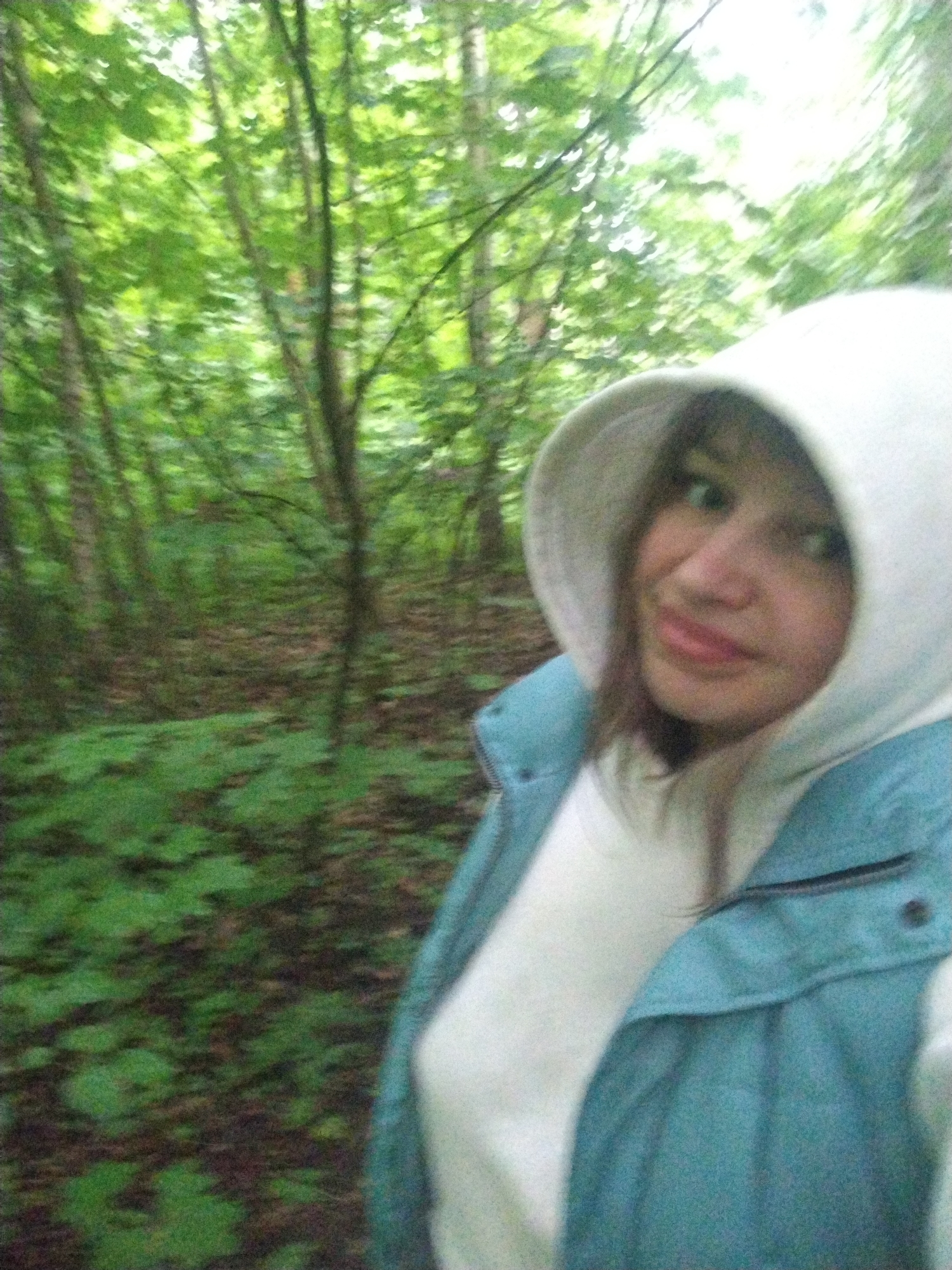 A blurry image shows a person wearing a white hooded sweatshirt and a blue jacket in a forest. The background consists of green trees and foliage, suggesting a wooded area. The person is likely located in the city of Rusanovo, Russia.