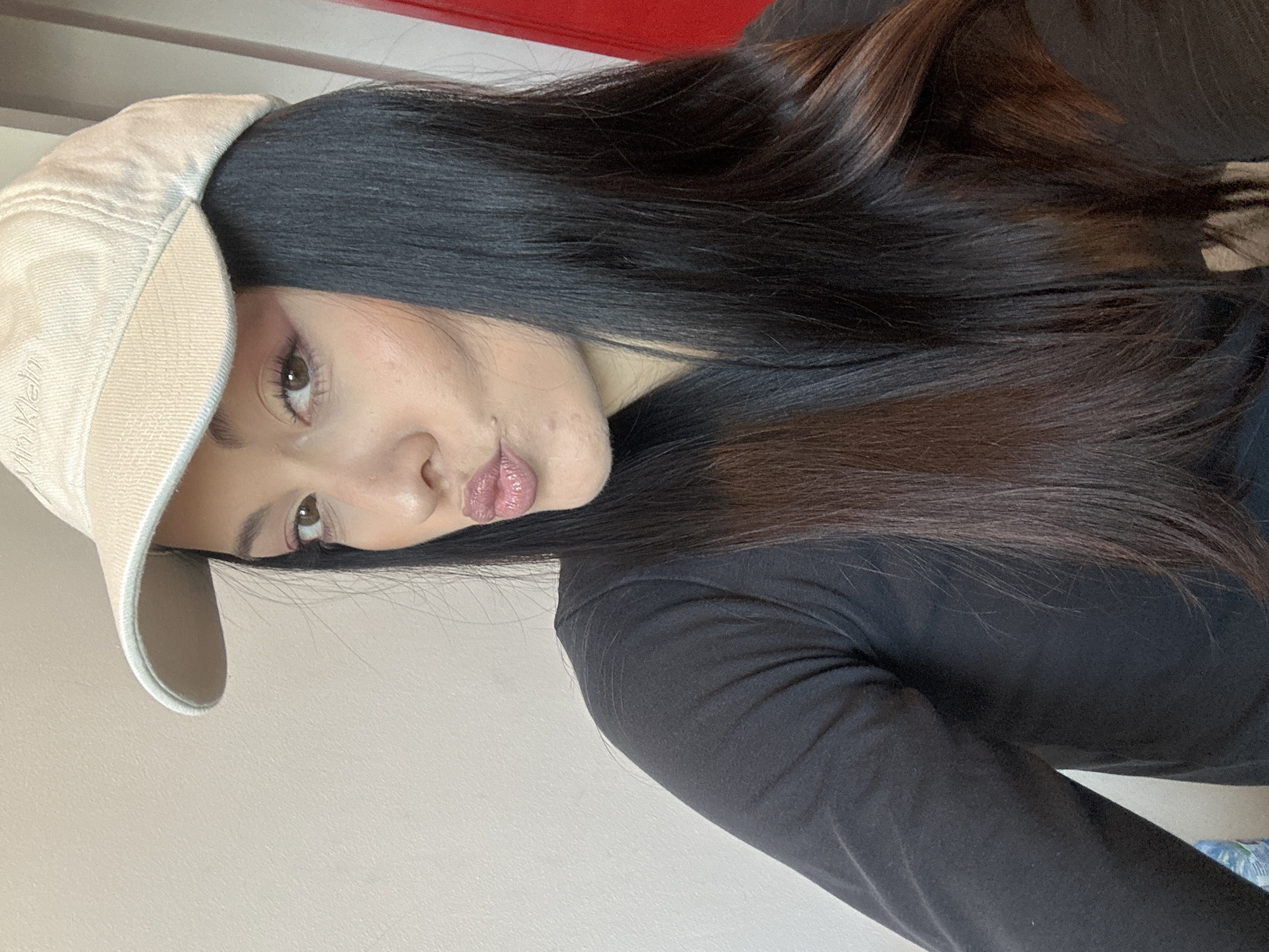 A young woman with dark hair and a beige baseball cap is facing the camera with pursed lips. She is wearing a long-sleeved black top. The background is a plain wall, with a hint of red color visible at the top. The photo is taken indoors and has a slight tilt. The location is not discernible from the provided image. Therefore, it is impossible to determine if this image was taken in Burgas, Bulgaria.