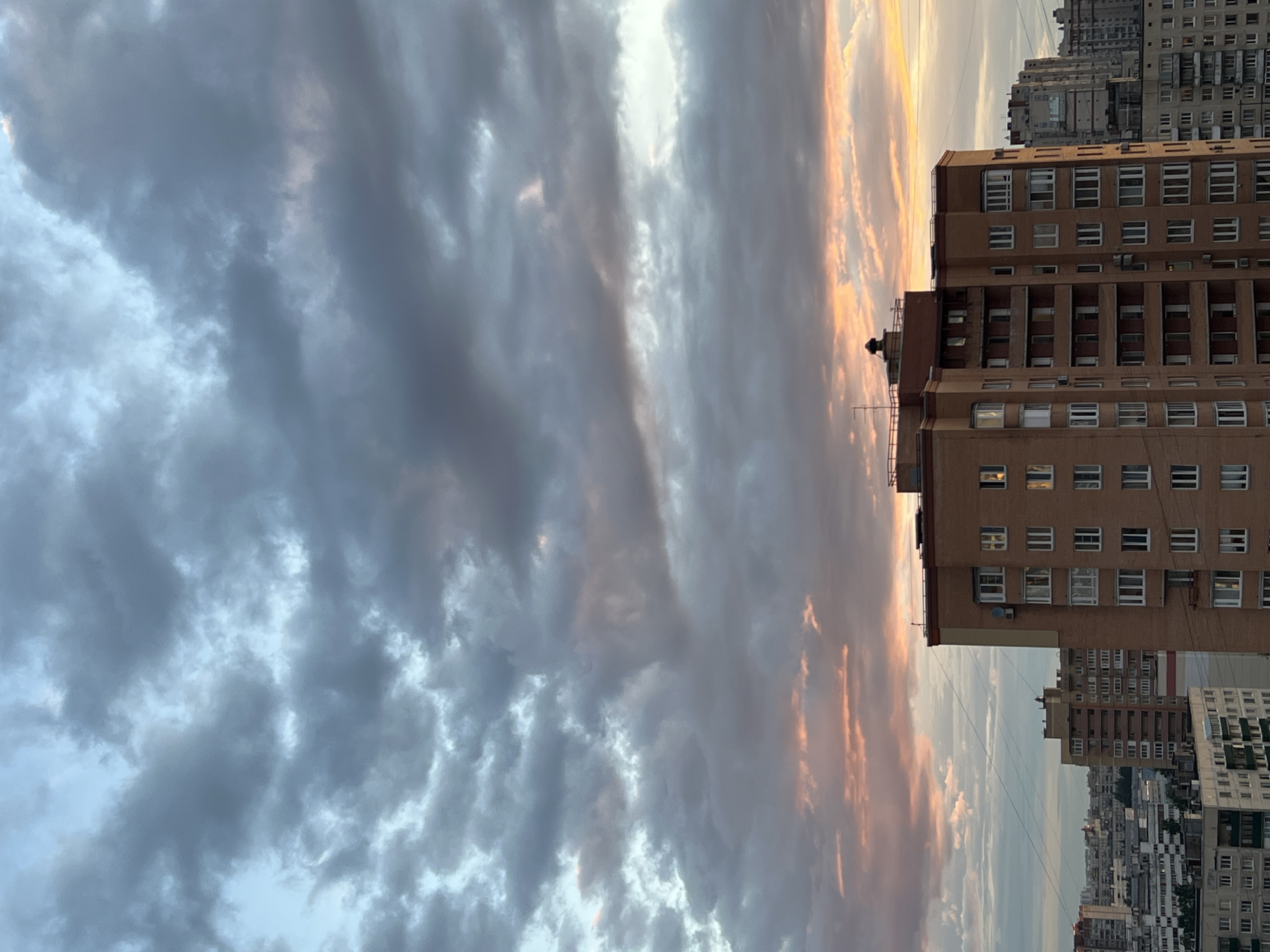 The image captures a city skyline at sunset, with a dramatic sky filled with clouds. The foreground is dominated by the side of a tall, multi-story building with a brown facade. The building features numerous windows, many of which appear to have lights on, suggesting it is residential or office space. Above this building, more buildings of varying heights and architectural styles can be seen receding into the distance, creating a dense urban landscape. The sky exhibits a gradient of colors, transitioning from deep grays and blues on the left to soft oranges and pinks on the right, indicative of the sunset. Wispy clouds stretch across the frame, catching the fading light and creating textured patterns in the sky. The overall scene evokes a sense of urban life against a natural spectacle, with the setting sun casting a warm glow over the city. No people or specific activities are clearly discernible. The time of day is clearly sunset. The weather appears to be partly cloudy. No visible text is present in the image.
