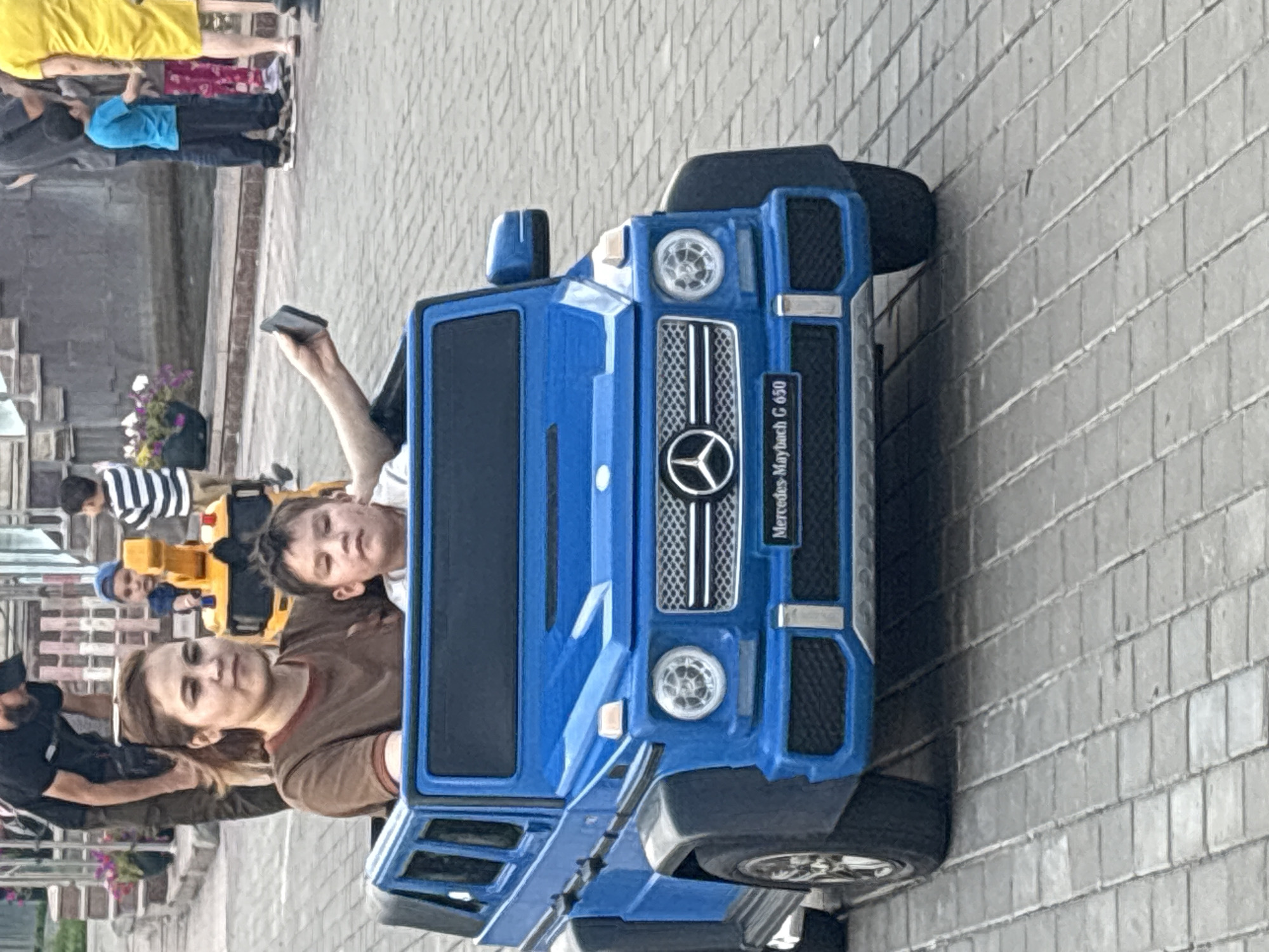 In Astrakhan, Russia, on a paved walkway, a woman with brown hair in a brown shirt is sitting in the driver's seat of a blue toy Mercedes-Maybach G 650. Next to her, a young boy with dark hair is in the passenger seat. The boy is holding a black mobile phone in his right hand and appears to be looking towards the camera. Behind them, a man in a black t-shirt and cap is visible, and further in the background, a yellow toy construction vehicle with a crane is parked. Several other people, including children and adults, are present in the background, some standing near a water feature. The overall scene depicts a recreational outing in a public urban space.