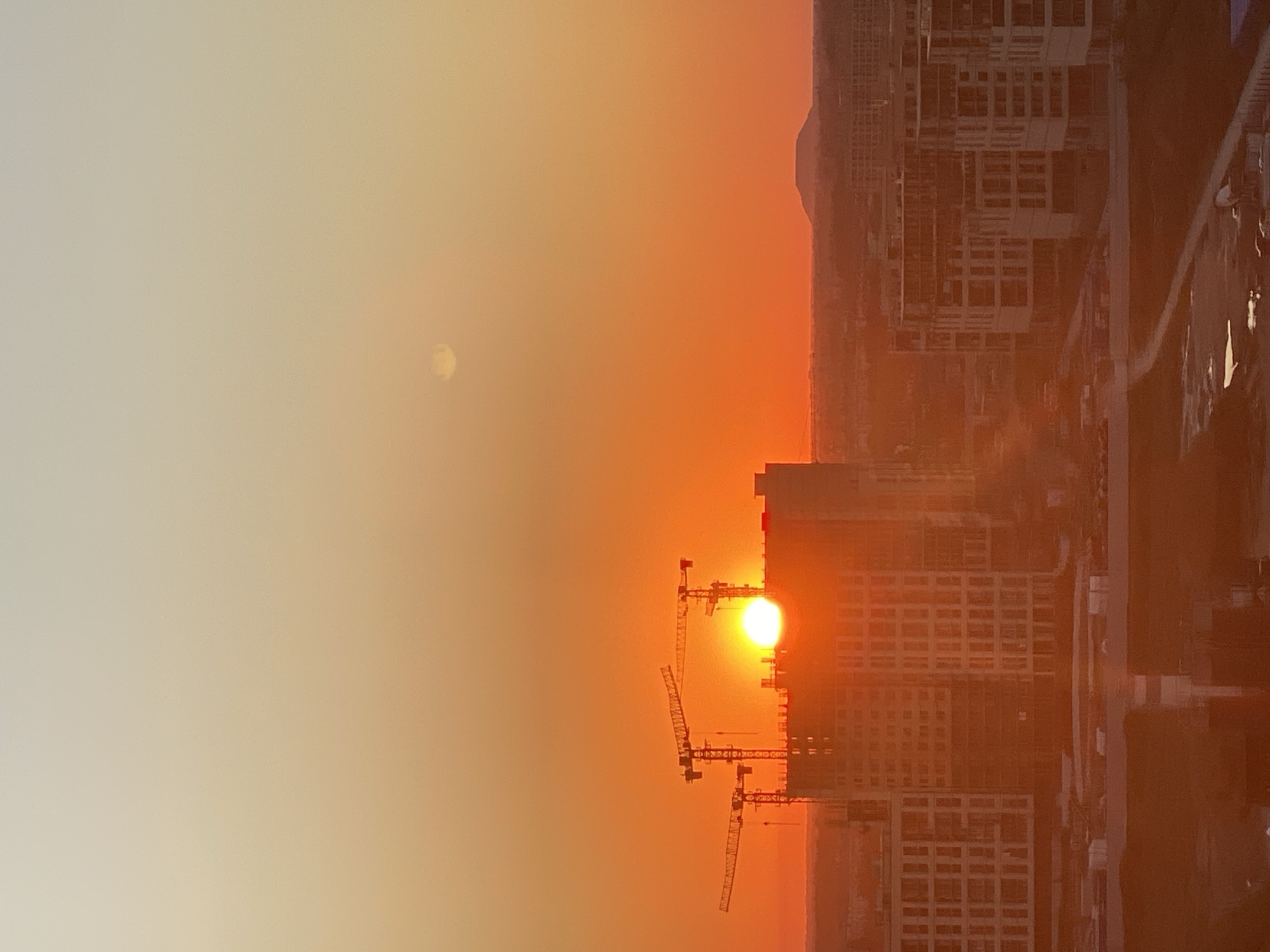 This image captures a vibrant sunset over the city of Novotitarovskaya, Russia. The sky is a brilliant gradient of orange and yellow, with the sun a bright, incandescent orb positioned just above the horizon. The sun is partially obscured by the silhouette of a large, modern building under construction, with several construction cranes extending into the sky. Multiple other buildings, some appearing to be apartment complexes, are visible in the middle ground, also appearing to be under construction or newly built. In the far distance, a conical hill or mountain is faintly visible against the hazy sky. The foreground shows a lower-lying urban landscape, with roads and some scattered elements that suggest ongoing development. The overall scene depicts a rapidly developing urban environment bathed in the warm, dramatic light of sunset.