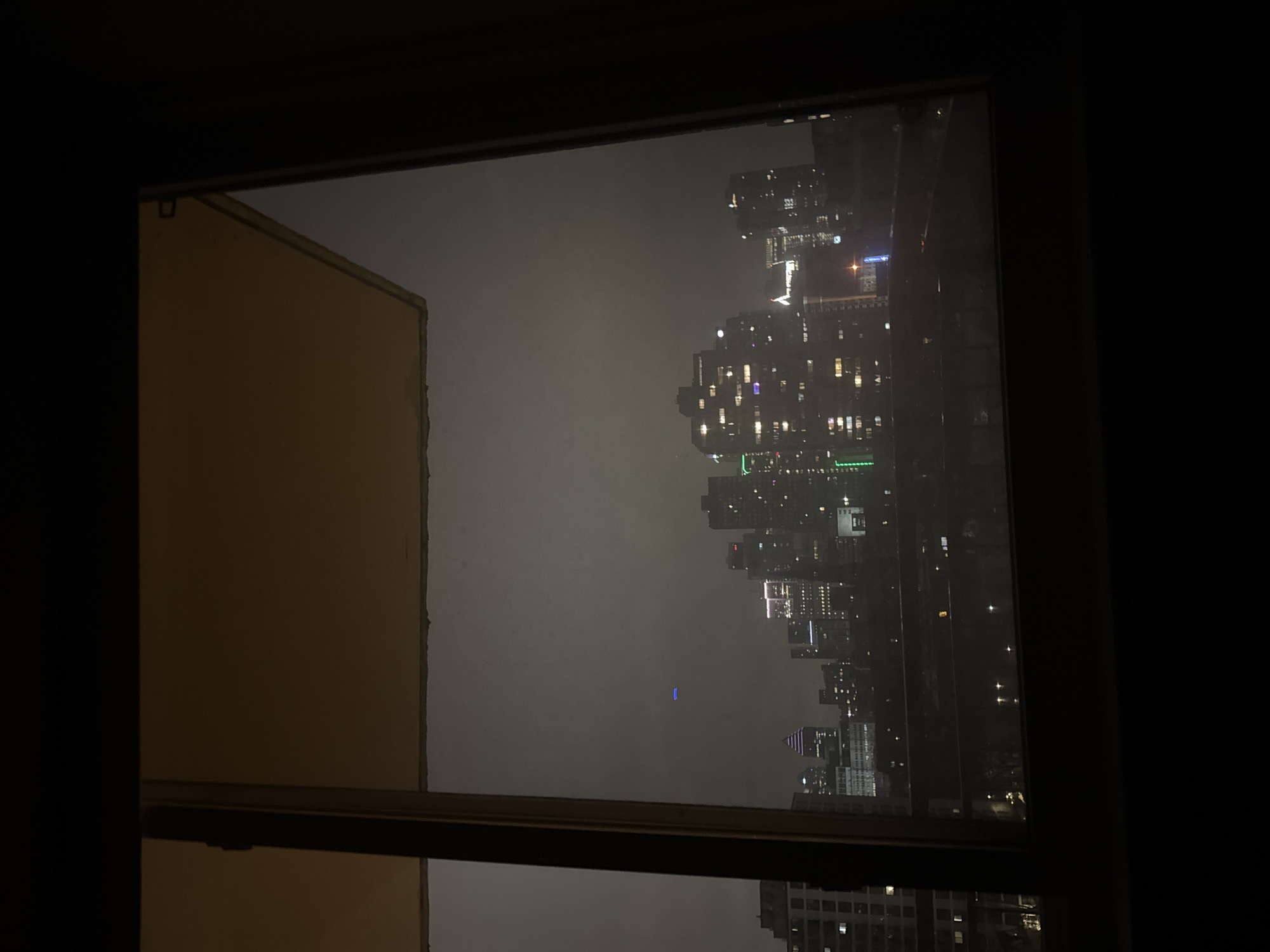 The image shows a view of a city skyline at night, seen through a window. The skyline consists of several high-rise buildings with lights visible in their windows. There is a hazy or foggy atmosphere obscuring some of the details of the cityscape. The buildings appear to be densely packed together. The window frame is visible in the foreground, with a portion of a building adjacent to the window on the left side of the frame. The rest of the room is dark.
The city is likely Montreal, Canada, given the architecture and skyline. More specifically, the view is of downtown Montreal from a high-rise building, likely near the city center.