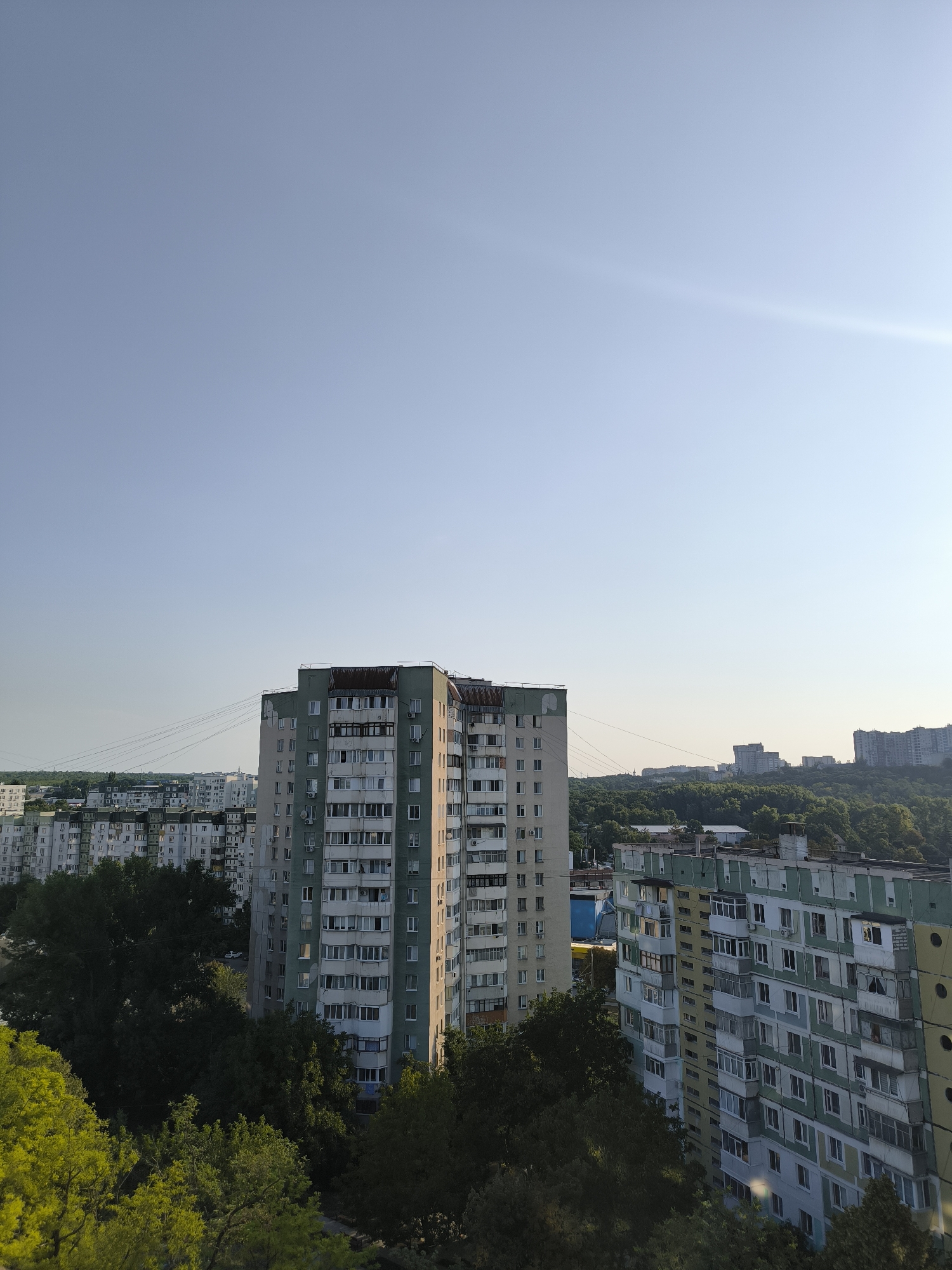 This is a medium-shot, high-angle, exterior, daylight photograph of a residential area in Chișinău, Moldova. In the foreground, on the right, is a multi-story apartment building painted in sections of light green, yellow, and light blue. The building has numerous balconies with glass enclosures and visible windows. To the left of this building, and slightly further back, is another tall apartment building, predominantly light gray with white window frames and balconies. In the midground, there are more apartment buildings of varying heights and colors, some appearing older and more uniformly gray. A dense canopy of green trees partially obscures the lower levels of the buildings. Power lines are visible stretching across the midground. In the background, more apartment buildings and structures can be seen on a hillside covered in trees, under a clear, pale blue sky with a faint sun flare on the right side of the frame. The scene depicts a typical urban residential landscape.