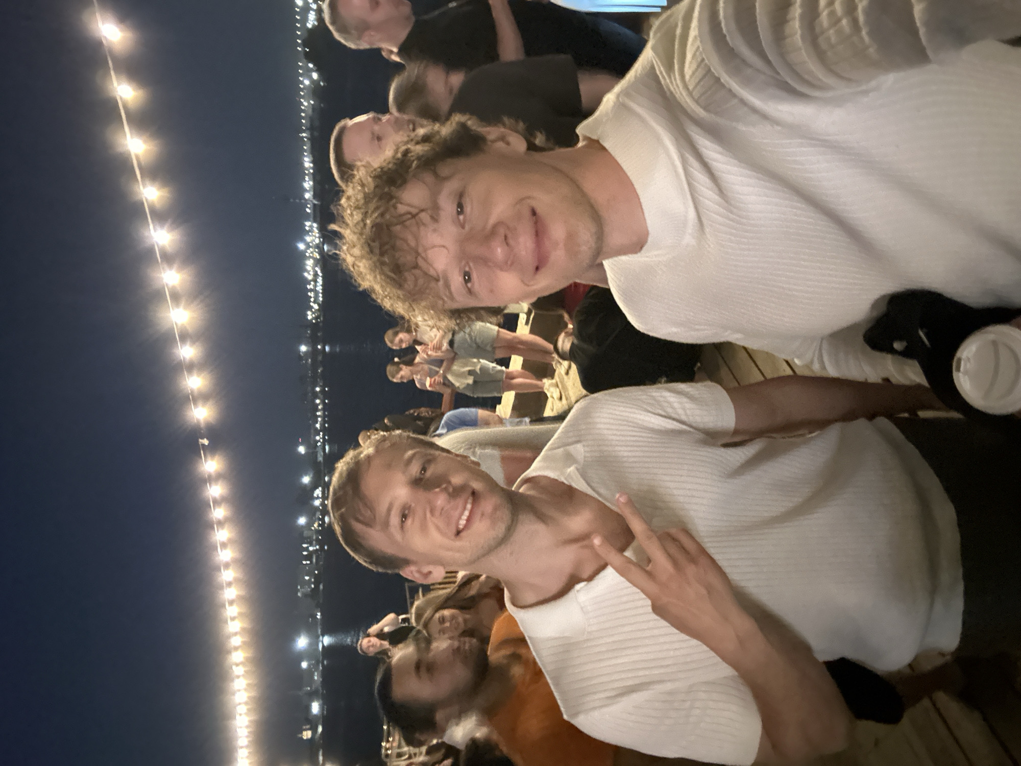 The image depicts a nighttime outdoor gathering, possibly a celebration or party, near a body of water. In the foreground, two young men are taking a selfie. The man on the left is wearing a white, short-sleeved, ribbed polo shirt and has his peace sign visible. The man on the right is wearing a white, long-sleeved, ribbed sweater and is holding a white paper cup. Both men are smiling at the camera.
In the background, a crowd of people are visible. They are casually dressed, suggesting a relaxed atmosphere. Some individuals are holding drinks. The environment appears to be a waterfront area, with lights from a city skyline visible across the water. String lights are illuminated overhead, casting a warm glow on the scene. The overall setting suggests a lively social event taking place in an urban environment at night. The presence of a prominent city skyline across the water indicates that this scene is located within the city of Saint Petersburg, Russia.