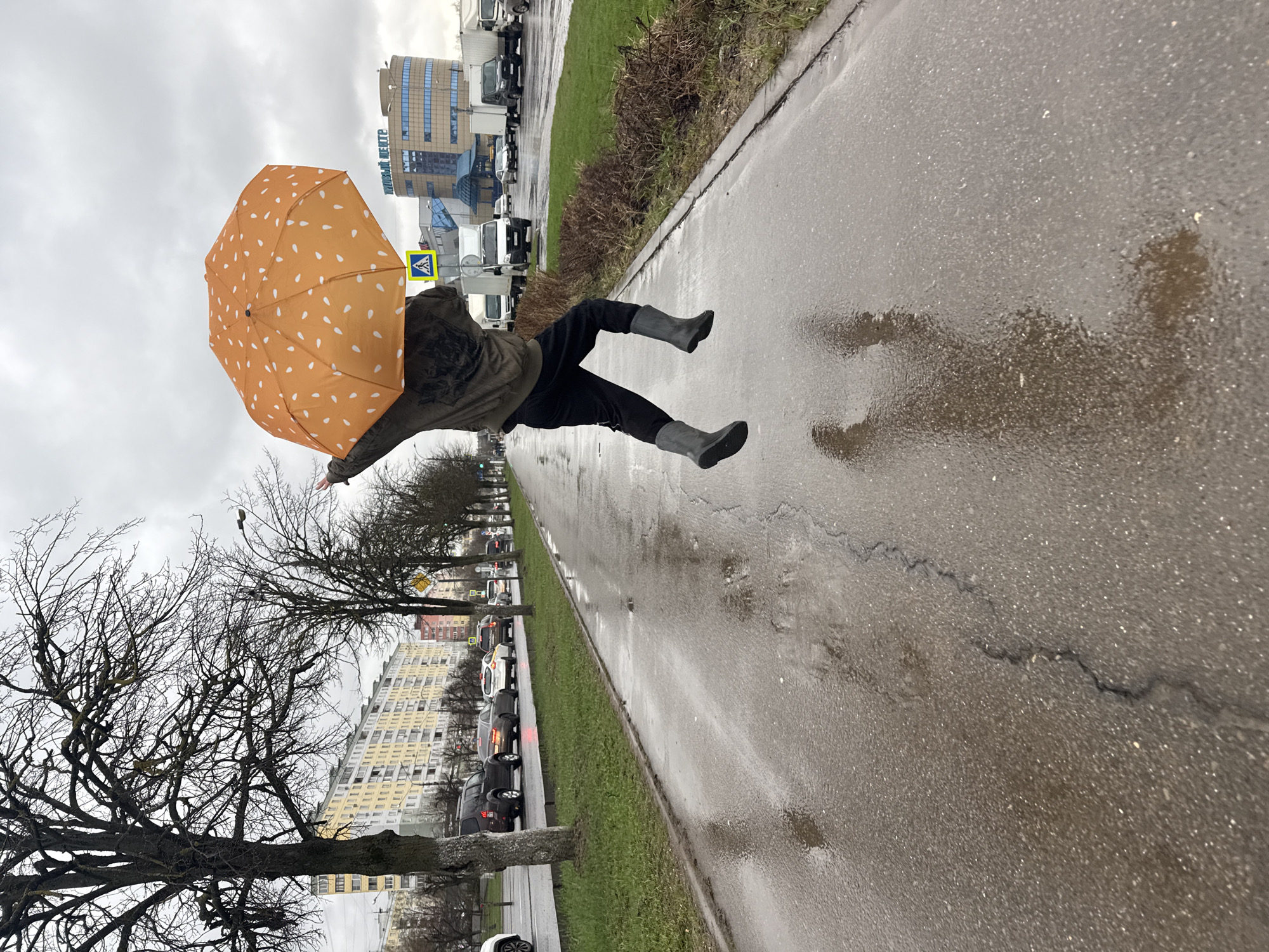 A person, presumably female, is captured mid-air, seemingly floating or falling, holding an orange umbrella with white speckles. They wear dark trousers, a dark-colored jacket, and light gray rubber boots. The setting appears to be a city sidewalk on a wet day, with reflections visible on the pavement. There's a line of parked cars along the side of the road and bare trees without leaves. In the background, there are buildings, including a modern glass and steel structure. Based on this, the photo appears to be taken in Dmitrov, Russia.