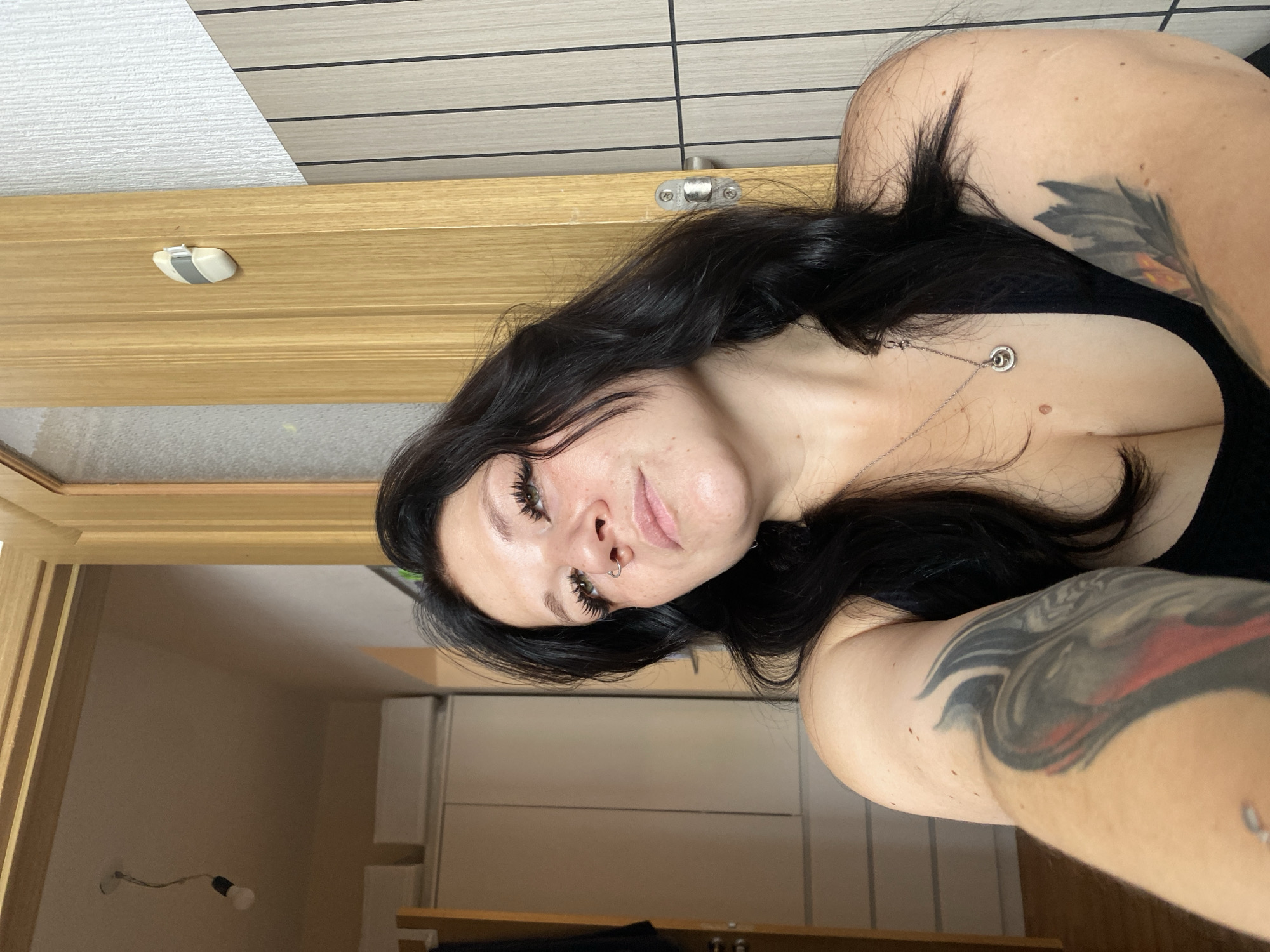 An indoor selfie captures a woman with long, dark, wavy hair and prominent false eyelashes looking upwards. She has a nose ring and a visible tattoo on her left arm, partially obscured by the camera's angle. Her right arm also shows a tattoo. She is wearing a black top and a silver pendant necklace. The background reveals a wooden doorframe, a white wall with vertical lines, and what appears to be a closet or wardrobe. A bare lightbulb hangs from the ceiling in the distance, and a small white object is attached to the doorframe. The overall setting suggests a residential interior. The location is identified as Unknown, Russia.