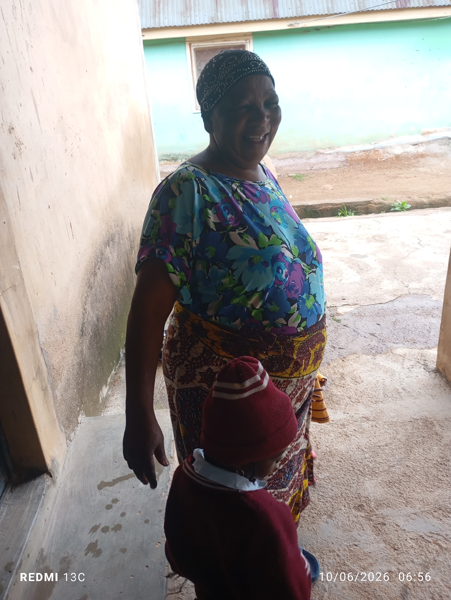 A full-shot photograph shows a Black woman standing outdoors and smiling while facing left. She is wearing a dark-colored headscarf, a brightly colored floral print top, and a patterned skirt. To the left of the woman is a beige wall, and to her right is an outdoor area with a concrete floor and a building with a teal-colored wall in the background. Partially visible in the foreground, on the lower right side of the image, is a child in a maroon sweater and matching beanie. The image is geotagged to an unknown location in Unknown, Unknown, and dated 10/06/2026 at 06:56.