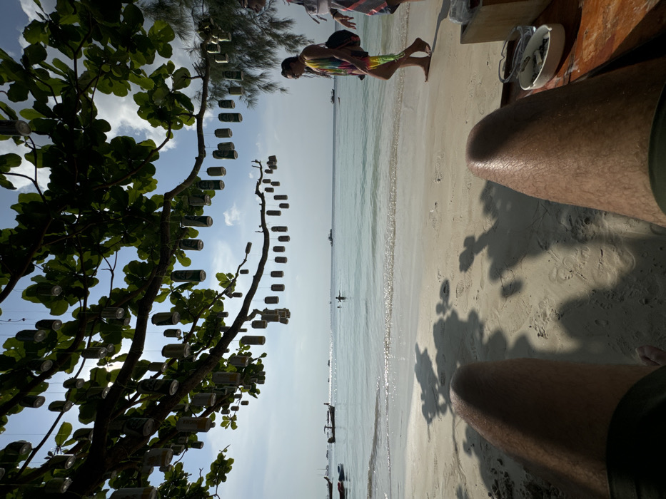 The image displays a first-person perspective of a beach in Unknown, Unknown. The observer's legs, clad in dark shorts, are visible in the lower foreground. To the right of the observer, a wooden surface holds a white ceramic ashtray containing cigarette butts and a small white dish. The scene transitions to a light sand beach and then to a light blue ocean with minor wave activity. A female subject, barefoot and wearing a multi-colored sarong and sunglasses, walks leftward along the waterline, carrying a dark shoulder bag. Her right arm is elevated. A second individual's torso, wearing a striped garment, is partially visible on the far right. In the ocean, one person wades or stands, with their head and shoulders above the surface. Multiple longtail boats and other vessels are present in the water. Overhead, a canopy of green tree leaves is present, from which numerous aluminum beverage cans, predominantly green and silver, are suspended by string. The sky is light blue with sparse white clouds, indicating bright daylight conditions.