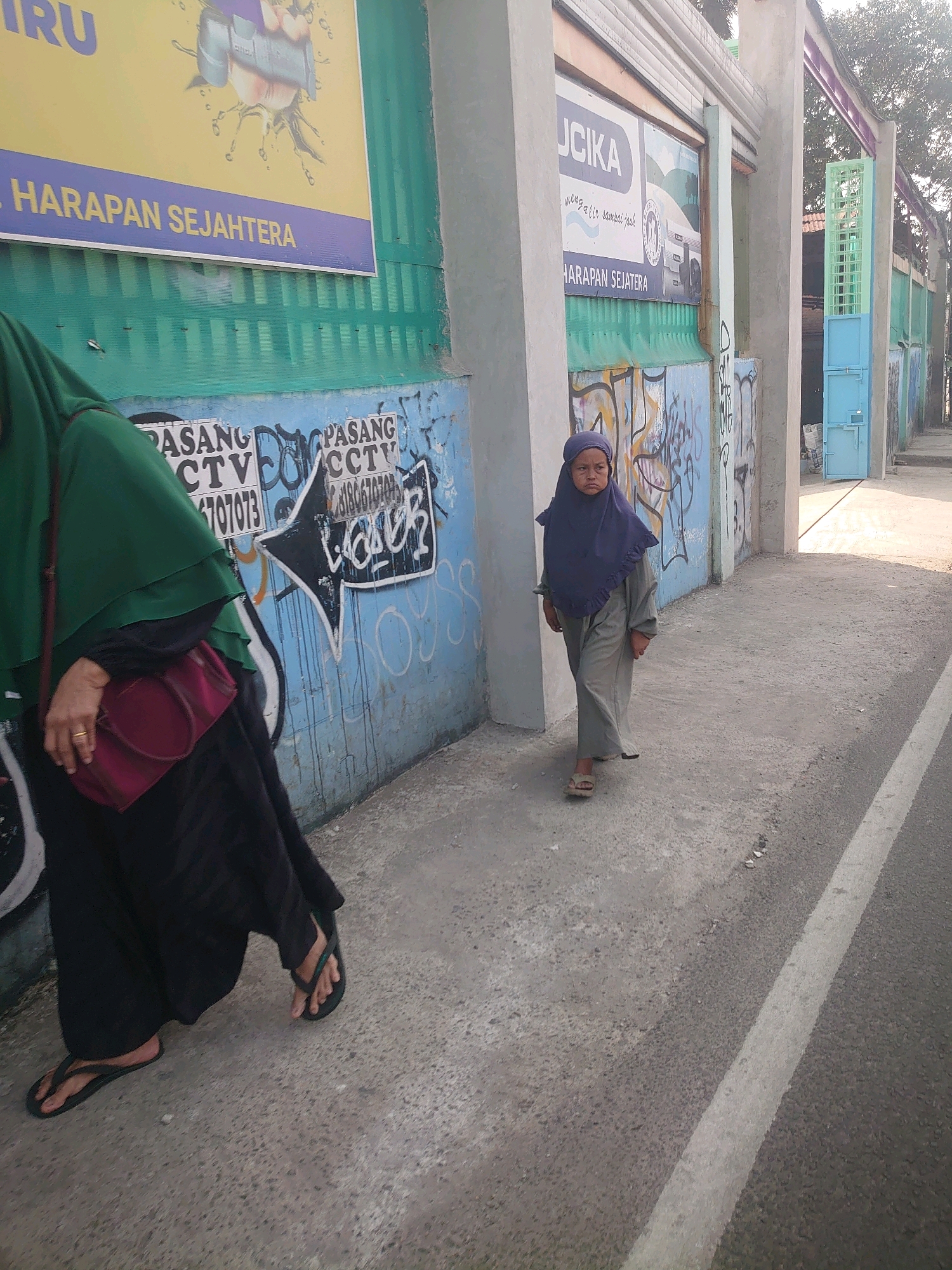 In Bojong Gede, Indonesia, a young girl wearing a dark blue hijab and a grey long-sleeved garment walks on a paved sidewalk. She is barefoot on her right foot and wearing a sandal on her left. Beside her, a person dressed in a green hijab and a black abaya, also wearing flip-flops, walks with a red purse slung over their shoulder. They are on a sidewalk next to a wall covered in graffiti and posters. The wall has posters with Indonesian text. Behind the subjects, a building entrance with a light blue door and a green metal gate is visible. The area appears to be outdoors, with sunlight casting shadows.
