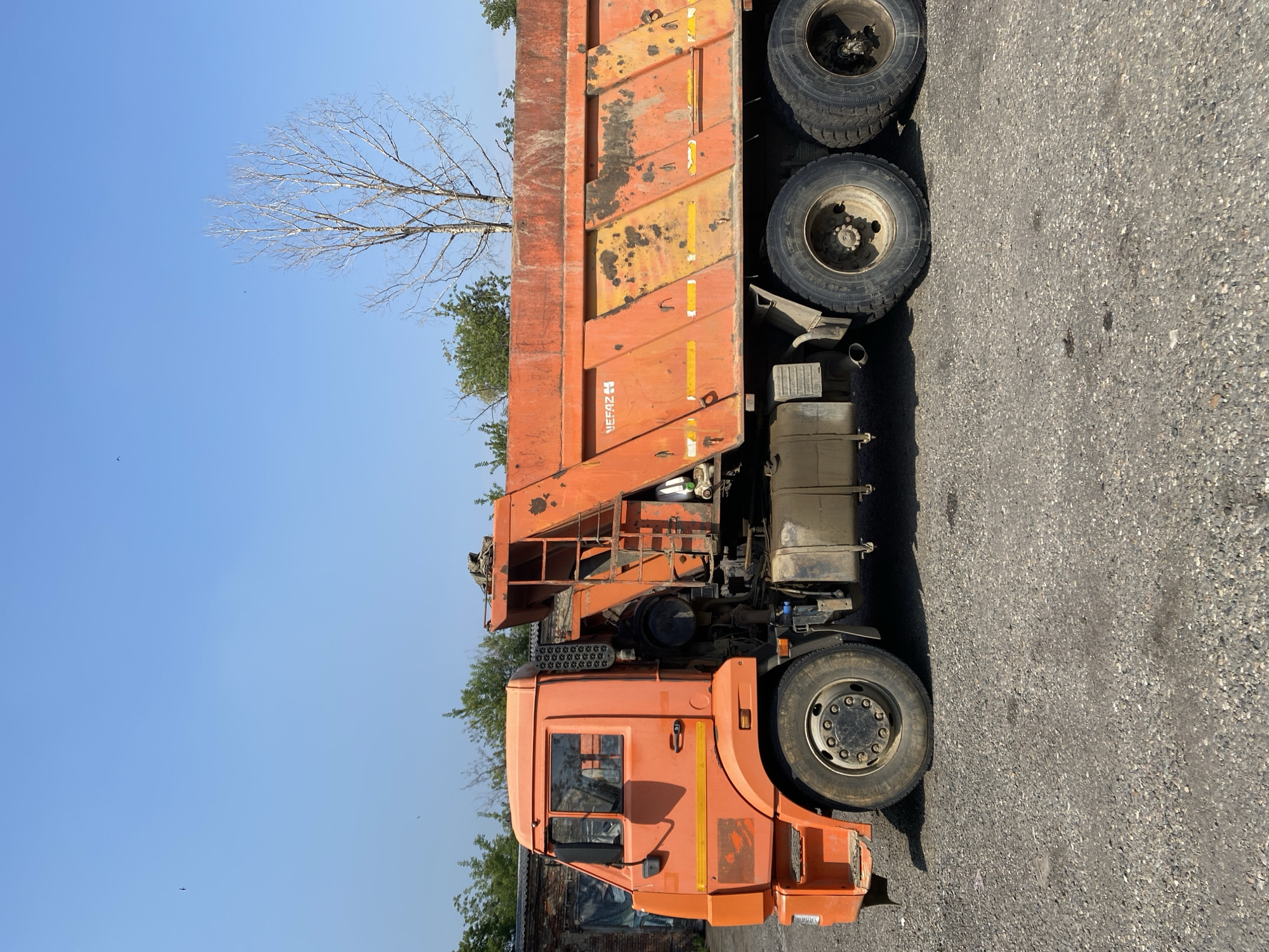 An orange dump truck, identified by the "NEFAZ" logo on its side, is parked on a gravel surface under a clear blue sky. The truck is facing left, with its front wheel visible on the left side of the frame and dual rear wheels on the right. The truck's bed is empty and shows signs of wear and tear, with rust and dirt accumulated in various areas. A ladder is attached to the side of the truck bed. In the background, a brick building with a garage door is visible on the left, and trees line the horizon. A leafless tree stands tall in the middle ground. The scene appears to be an industrial or construction yard. The image is dated as taken in Novokuznetsk, Russia.