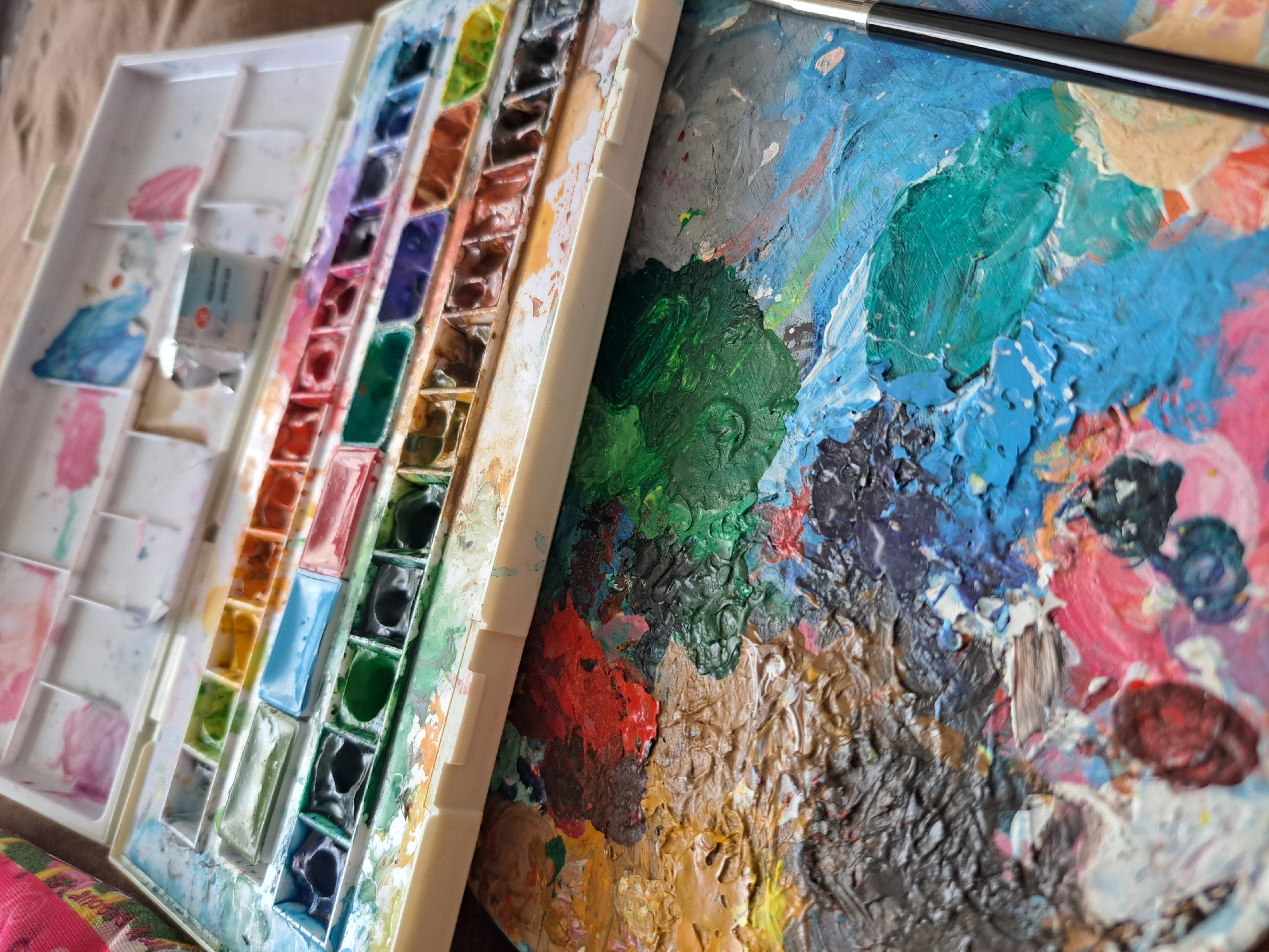 This close-up, high-angle shot showcases a watercolor paint palette and a separate mixing palette heavily laden with colorful, textured paint. The subjects are art supplies, specifically a white plastic watercolor paint box containing numerous rectangular pans filled with dried watercolors in a variety of hues, including blues, greens, purples, oranges, and browns. Adjacent to this is a larger, off-white mixing palette with a textured surface, where wet paints have been blended. The mixing palette displays thick impasto strokes of colors like vibrant blue, deep green, red, black, and various earthy tones, indicating recent use. A black-handled paintbrush lies across the top of the mixing palette, its bristles slightly smeared with paint. The background appears to be a textured, neutral surface, possibly fabric or a tablecloth, in a warm, muted tone. The lighting suggests an indoor setting, possibly a studio or workspace, with soft illumination. There is no discernible time of day or weather indicated. The overall mood conveyed is one of artistic creation and the tangible evidence of a painting process. No visible text is present in the image.
