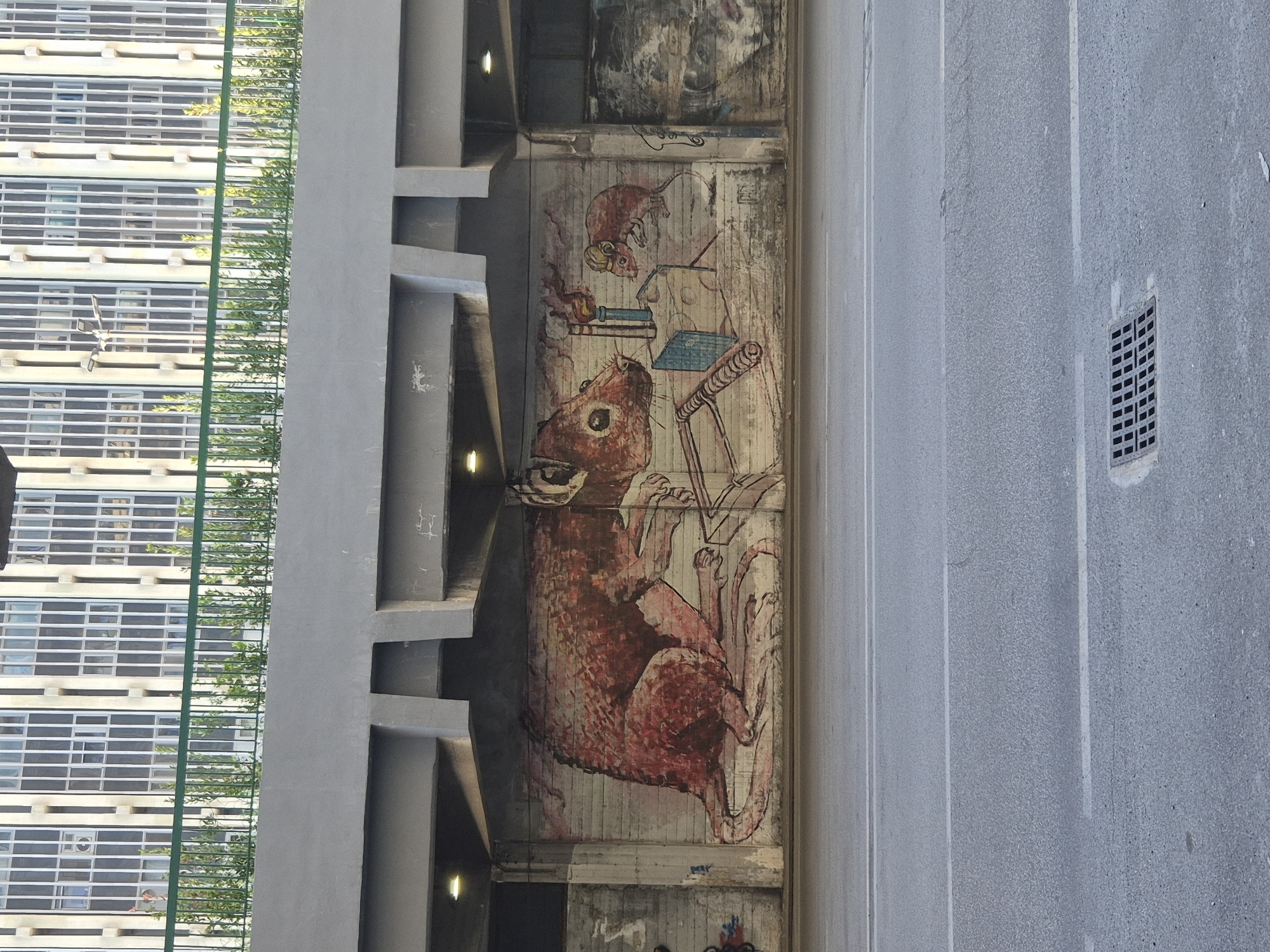 The image shows a mural on a concrete wall under an overpass in Taranto, Italy. The mural depicts two mice, one large and one small, interacting with what appears to be a piece of cheese and a device. The large mouse is on the left, facing right, with its body taking up a significant portion of the mural. The smaller mouse is to the right of the large one, also facing right, and appears to be working on something. The wall itself is brickwork, and the mural is rendered in a reddish-brown and white color palette. Above the mural, the overpass structure is visible, with exposed concrete and lighting fixtures. The overpass is supported by concrete pillars. In the background, a building with numerous windows is visible, suggesting an urban setting. The foreground shows a street with asphalt and white lane markings, and a drain grate.