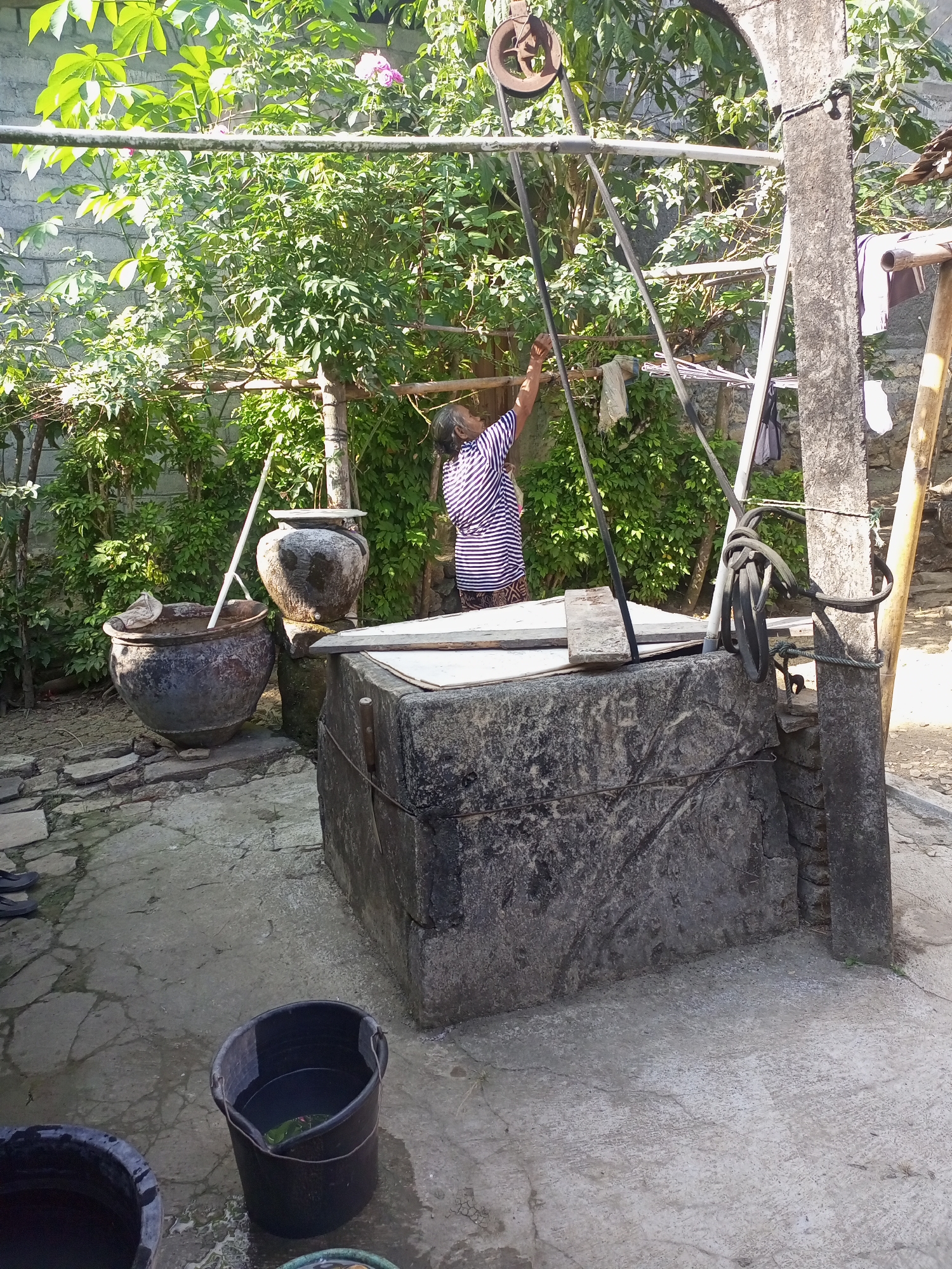 The image depicts an outdoor scene at a traditional well located in Nanggulan, Indonesia. A woman, wearing a striped shirt and skirt, is actively drawing water from the well using a pulley system. The well itself is constructed from large stone blocks and is covered with a wooden plank. To the left of the well, there are two large clay pots, one of which is empty and the other filled with water and a cloth. Buckets are also visible on the ground, one filled with water. The background is lush with greenery, including a dense wall of bushes and trees, suggesting a rural or semi-rural setting. Clothes are hanging on a clothesline to the right of the well, indicating domestic activity. The overall atmosphere is one of daily life and resourcefulness in a tropical environment.