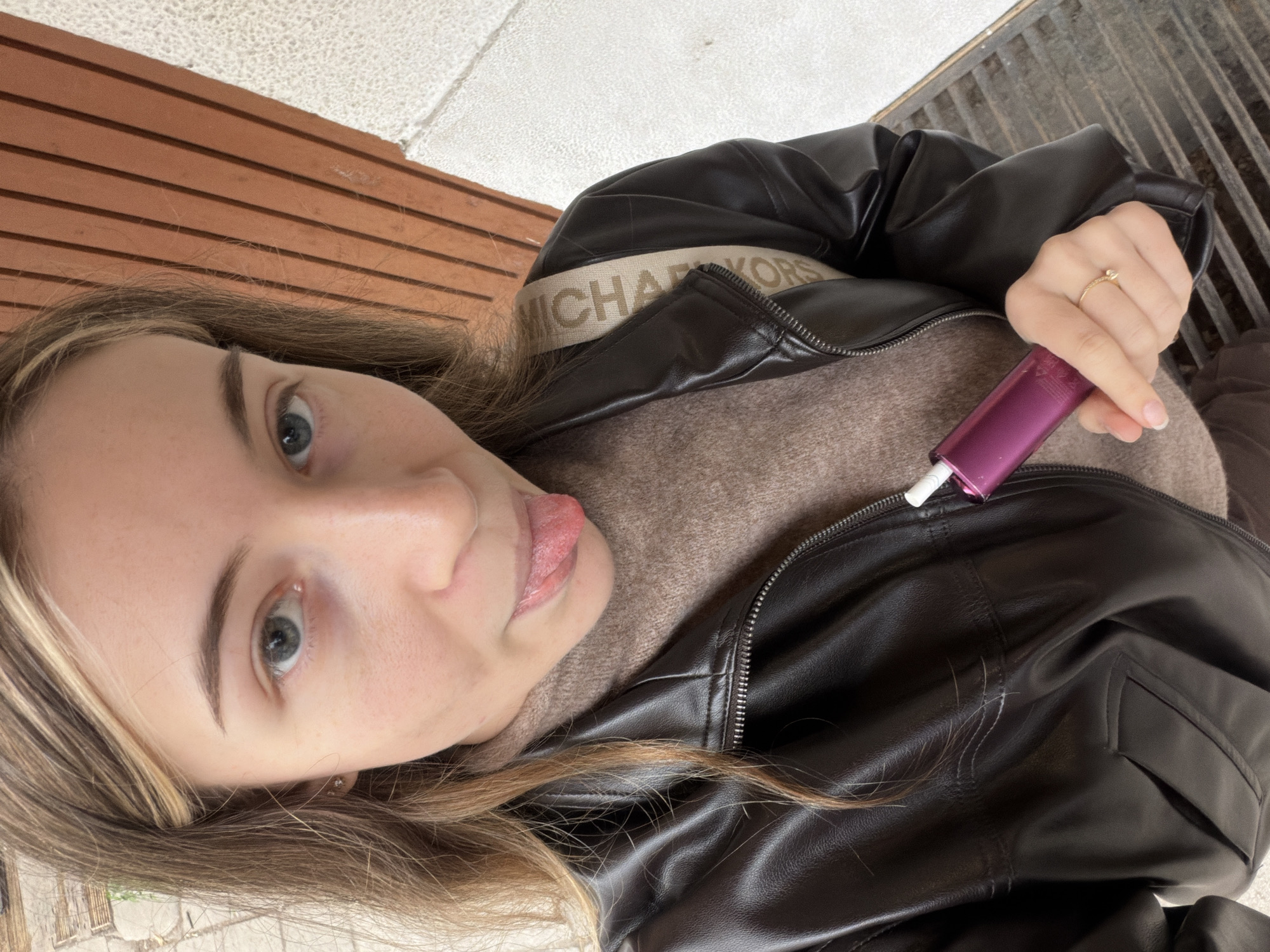 A woman with long blonde hair and blue eyes is shown from a slightly elevated, off-center perspective. She is wearing a black leather jacket over a light brown sweater with the word "MICHAEL KORS" visible on the collar. In her right hand, she holds a purple vape pen. Her tongue is slightly protruding from her mouth. She is sitting on a wooden bench. The background includes a light-colored tile surface and a gray metal grate. The location is identified as being in Plovdiv, Bulgaria.