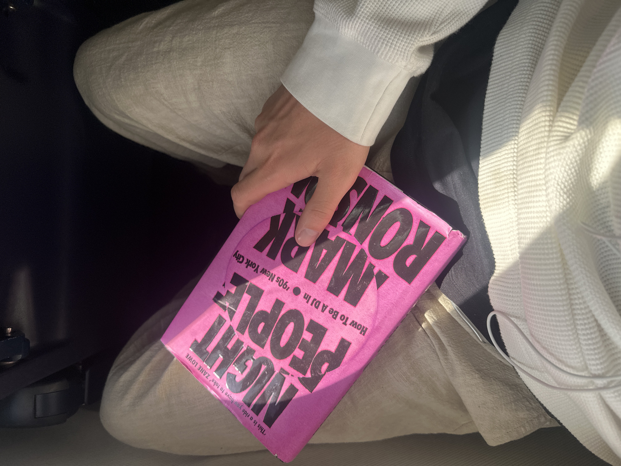 A person is holding a pink book in their lap. The book is titled "NIGHT PEOPLE: How To Be A DJ In '90s New York City" by MARK ROMV. The person is wearing light-colored pants and a white long-sleeved shirt. The book is held in the person's left hand. The setting appears to be indoors. The location is not within Braintree, United Kingdom, as the book title refers to New York City.